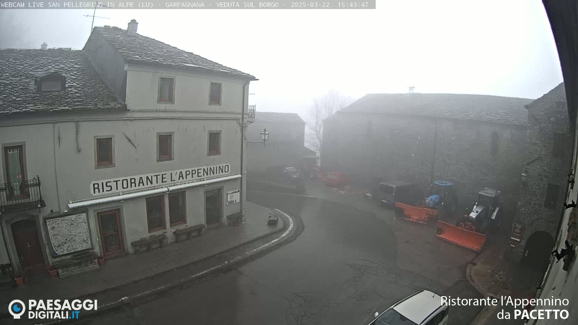 A foggy village square is seen, with snow plows parked near stone buildings, one of which is a restaurant.