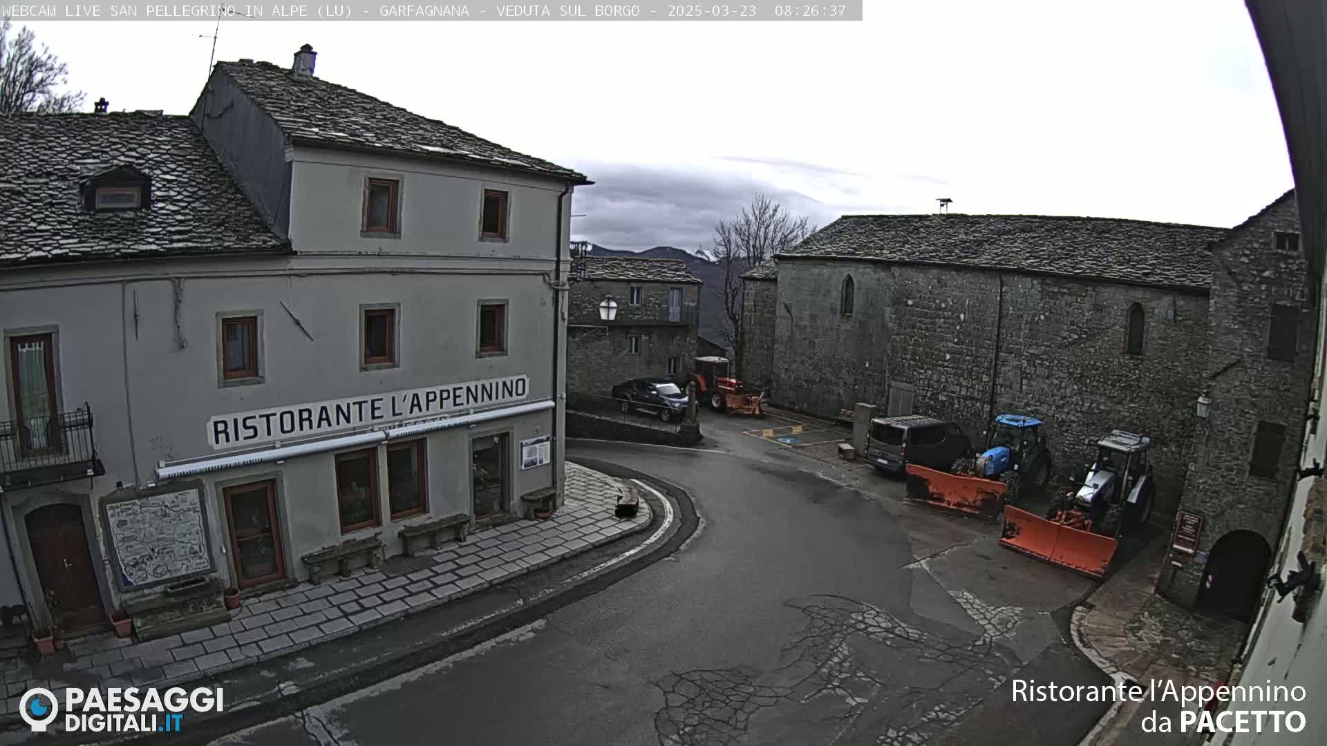 A village square with several stone buildings, some vehicles including tractors and snowplows, and overcast skies.