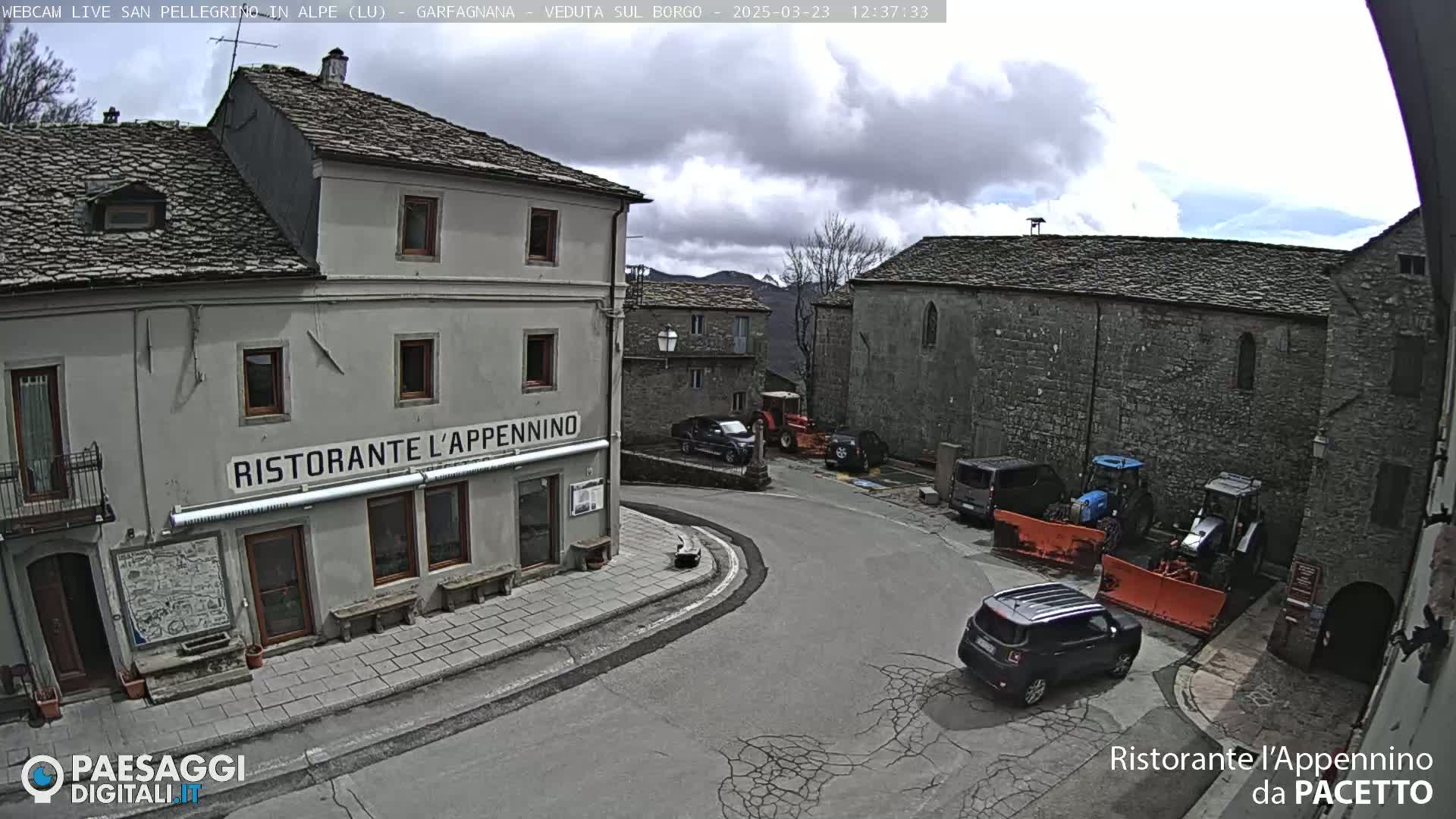 A village square with stone buildings, several parked cars and snowplows, under a partly cloudy sky.