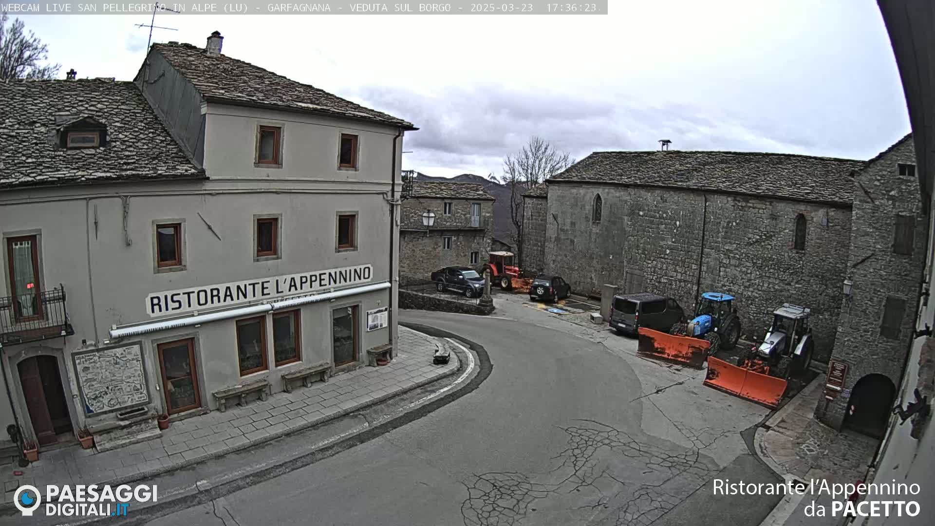 A village square with several stone buildings, some parked vehicles, and two tractors with snow plows, under a cloudy sky.