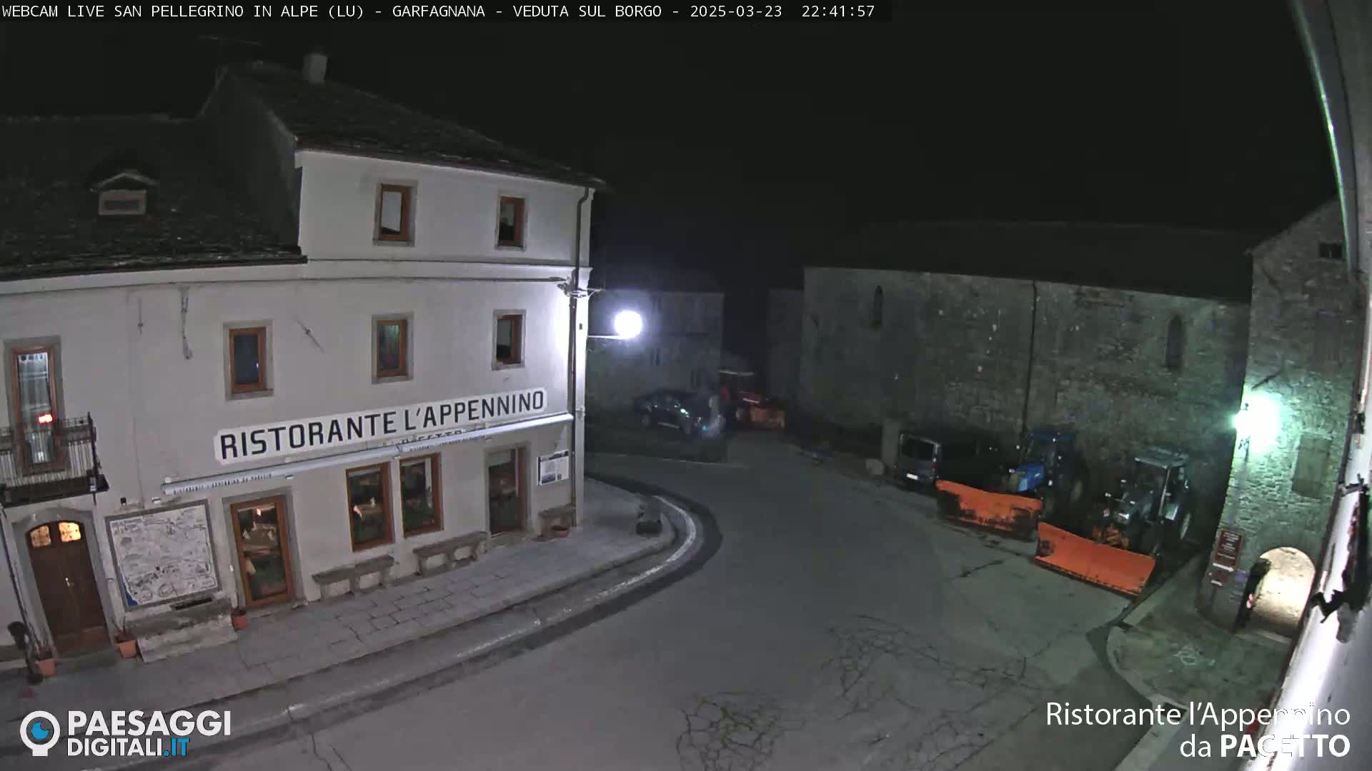 A nighttime view of a town square with a restaurant, several parked vehicles, and snowplows.