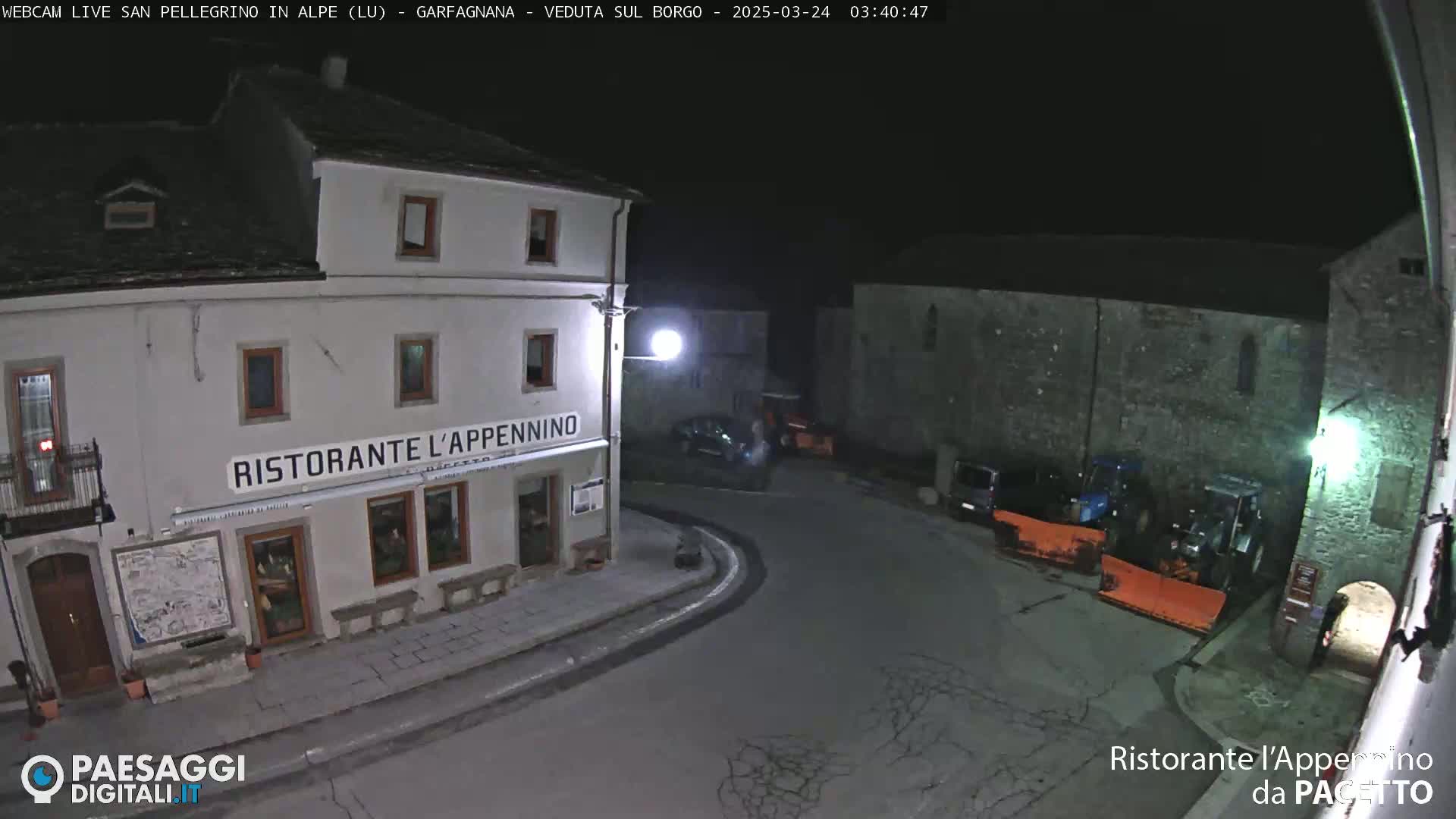 A nighttime view of a village square with a restaurant, several parked cars, and snowplows.
