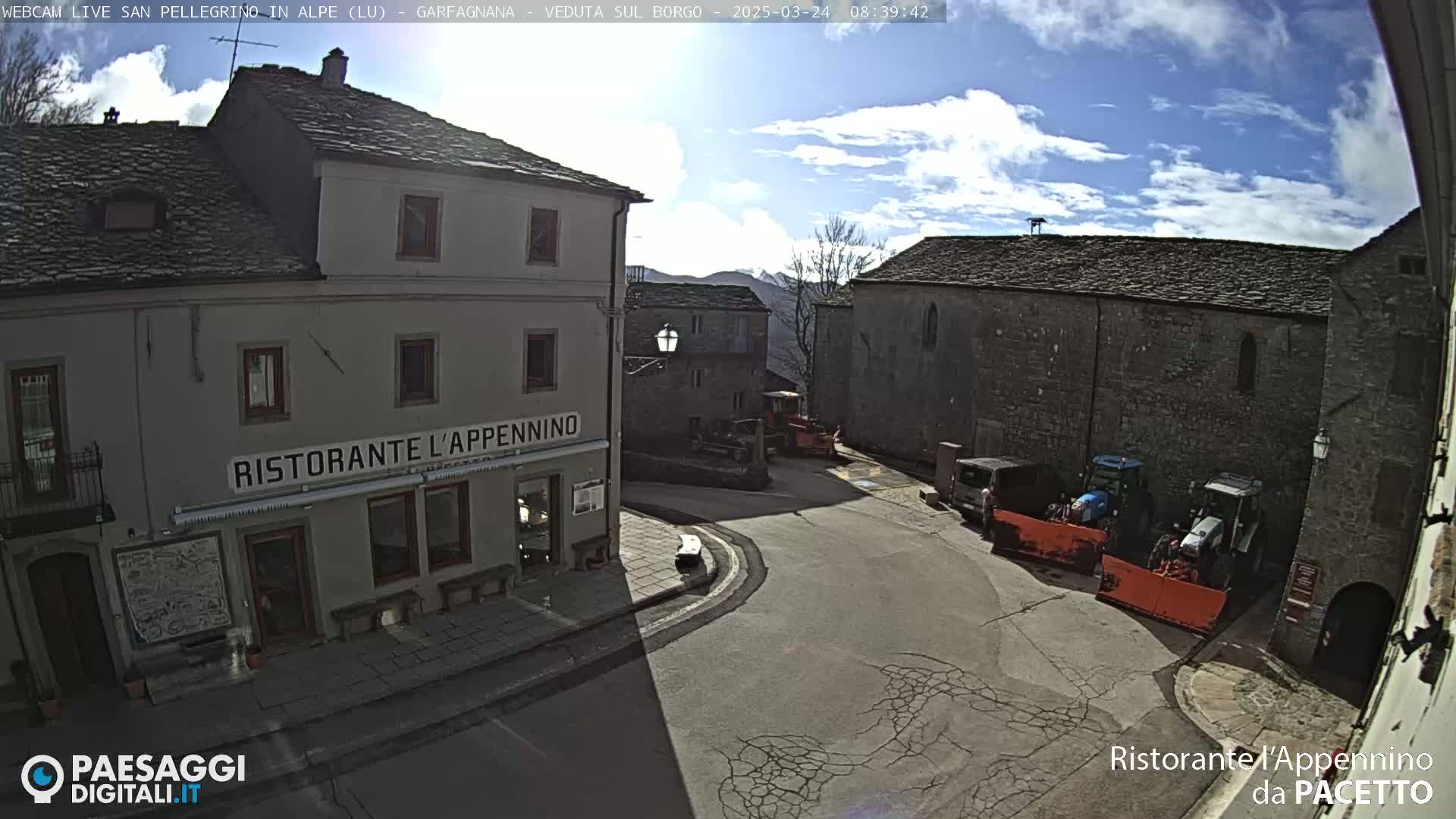 A sunny day reveals a village square with stone buildings, one of which is a restaurant, and several parked tractors and snowplows.
