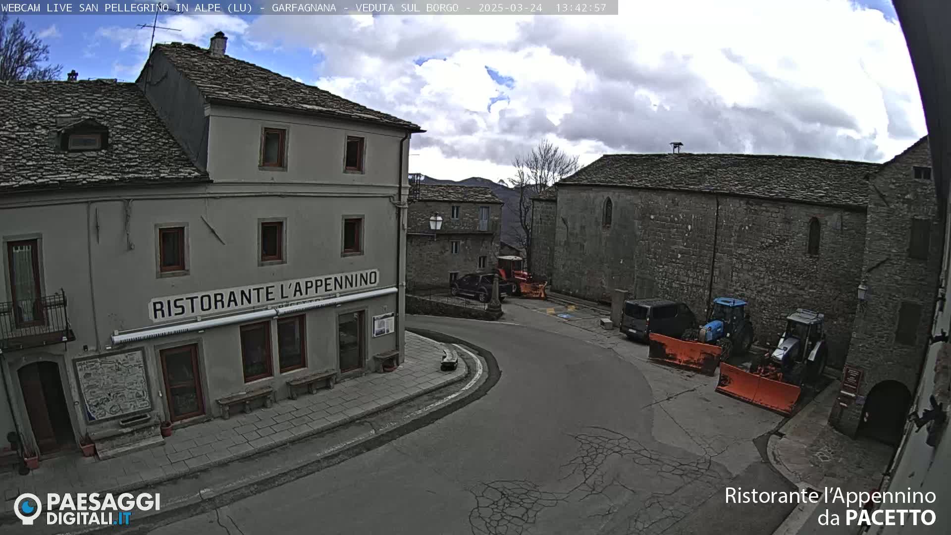 A partly cloudy day reveals a village square with stone buildings, a restaurant, and parked vehicles, including some snowplows.
