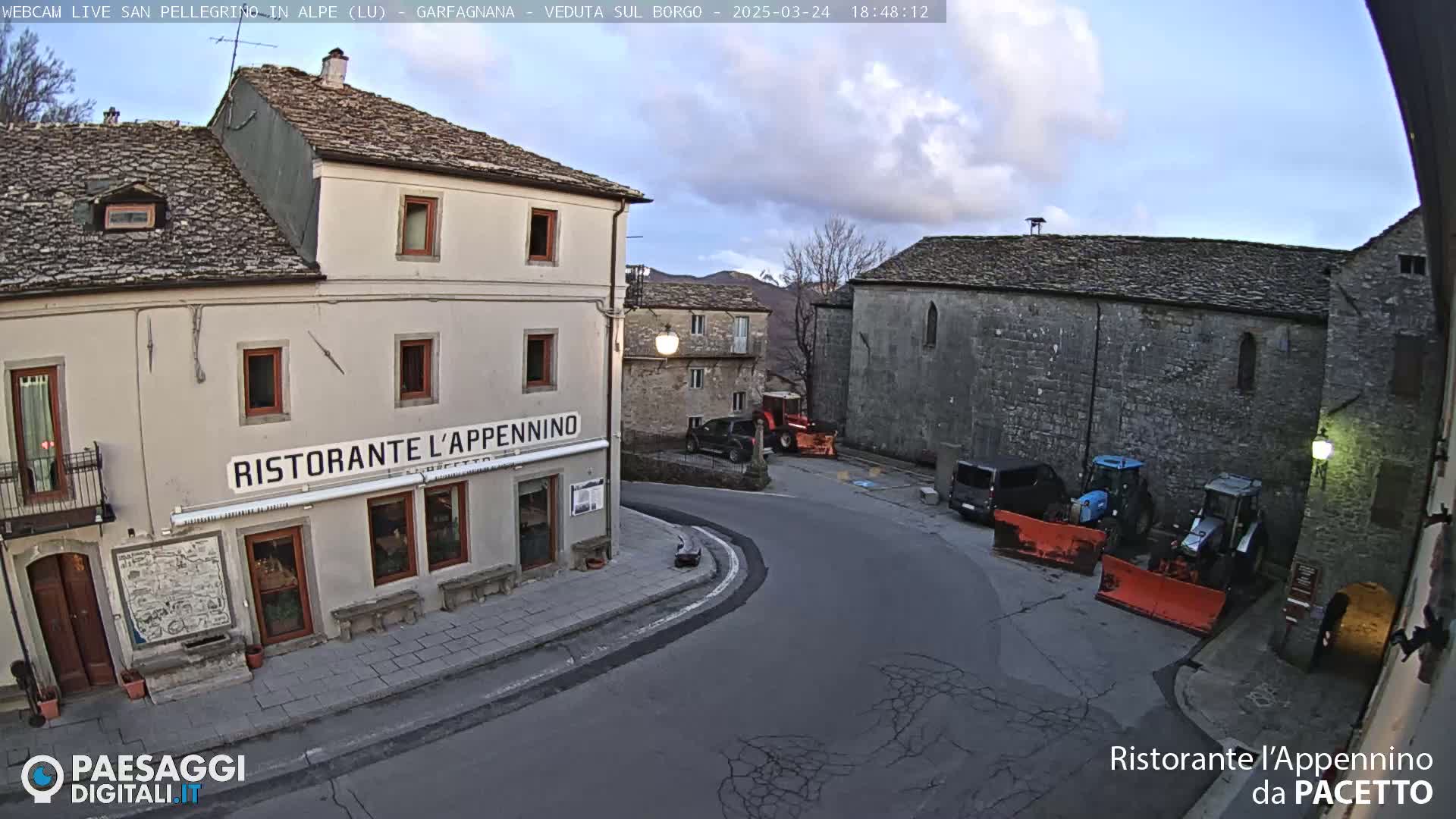 A street corner in a small town, under a partly cloudy sky, shows stone buildings, a restaurant, parked vehicles, and two tractors with snow plows.