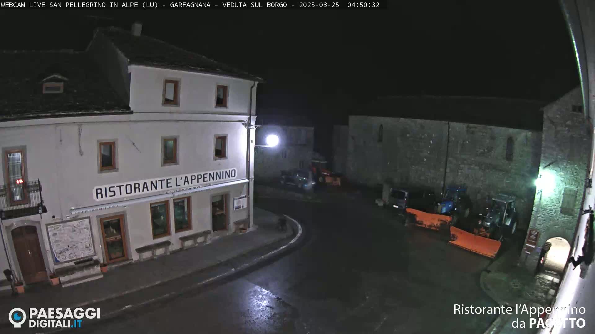 A nighttime view of a town square, featuring a restaurant and several parked vehicles including snow plows, under a clear, dark sky.