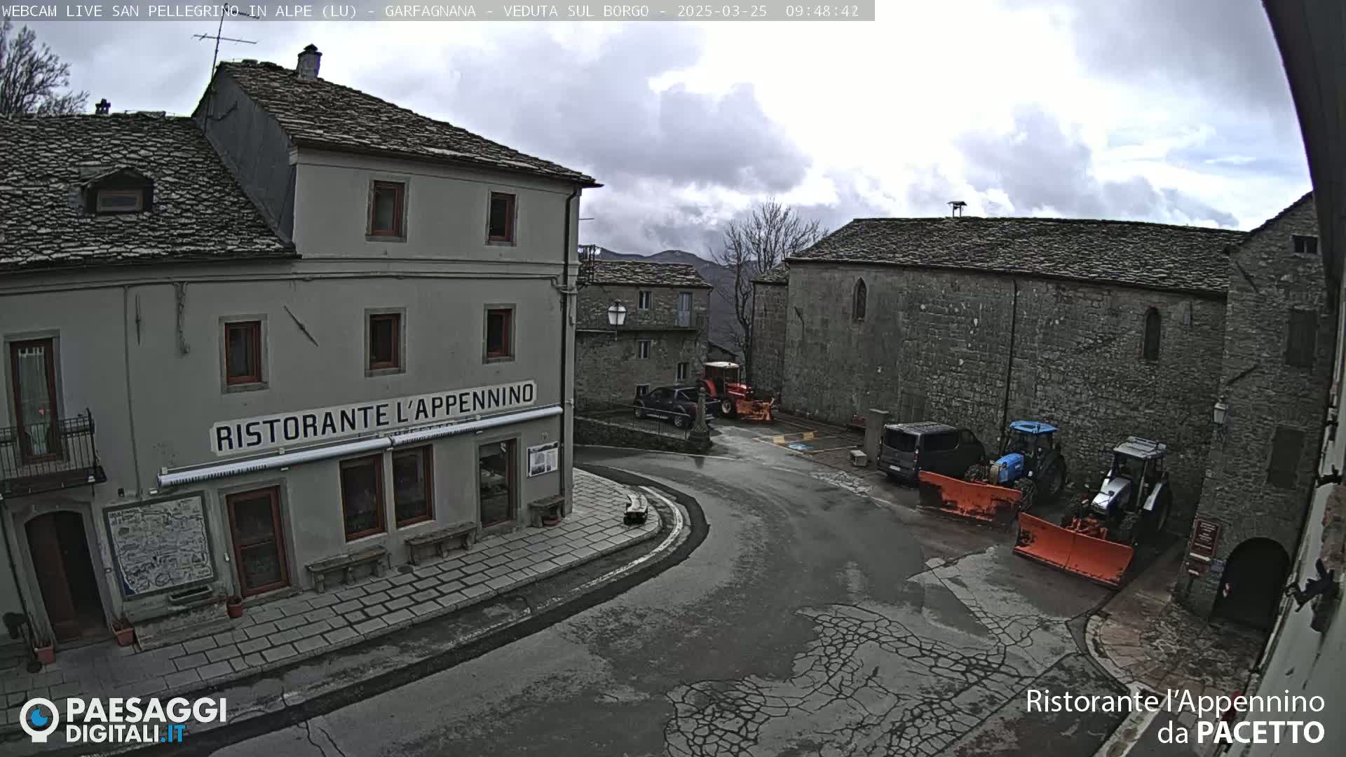 A stone village square with overcast skies, several vehicles, and snow plows are visible; a restaurant is prominent in the foreground.