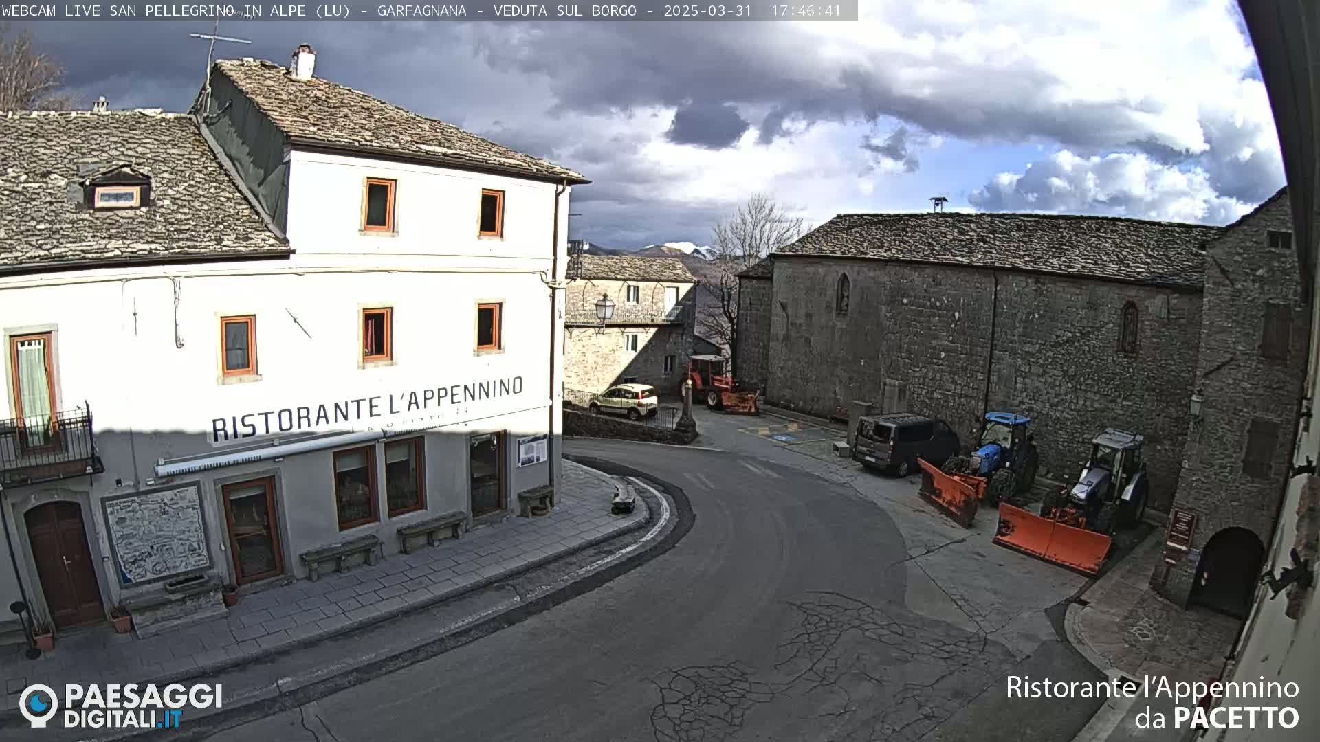 A partly cloudy day reveals a town square with stone buildings, including a restaurant, a few parked cars and vans, and snow-removal equipment.