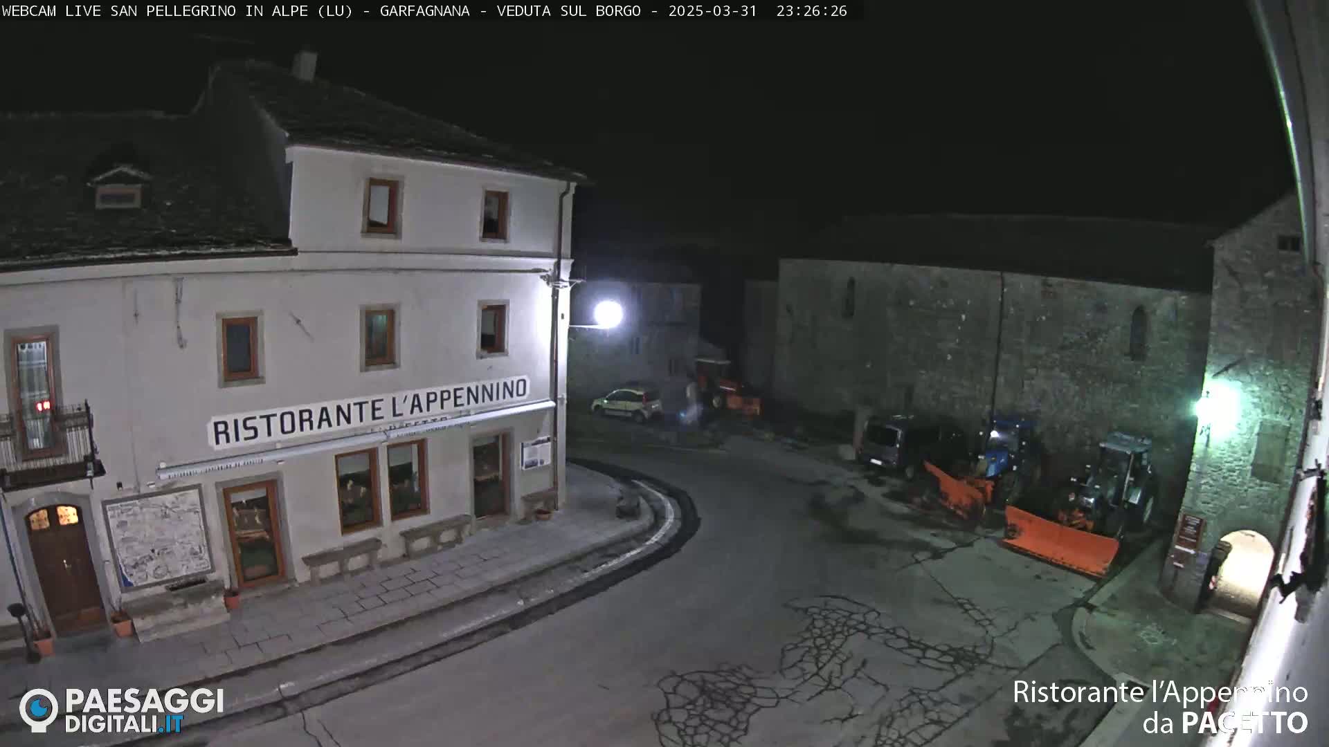 A nighttime, high-angle view of a town square with a restaurant, some parked cars, and snowplows.
