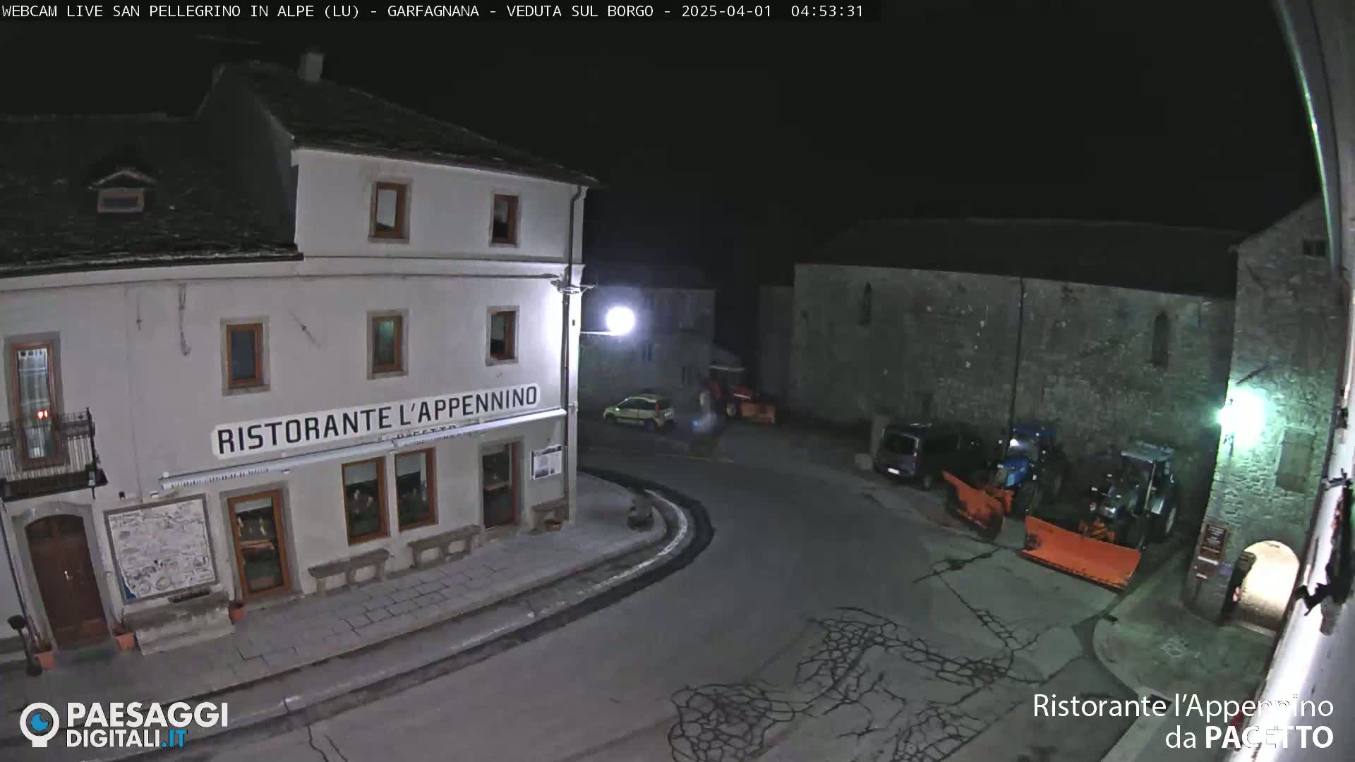 A nighttime view of a town square with a restaurant, a few parked cars, and snowplows.