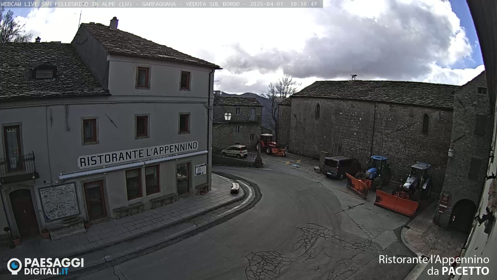 A partially cloudy day reveals a village square with stone buildings, a restaurant, several parked vehicles, and tractors with snow plows.