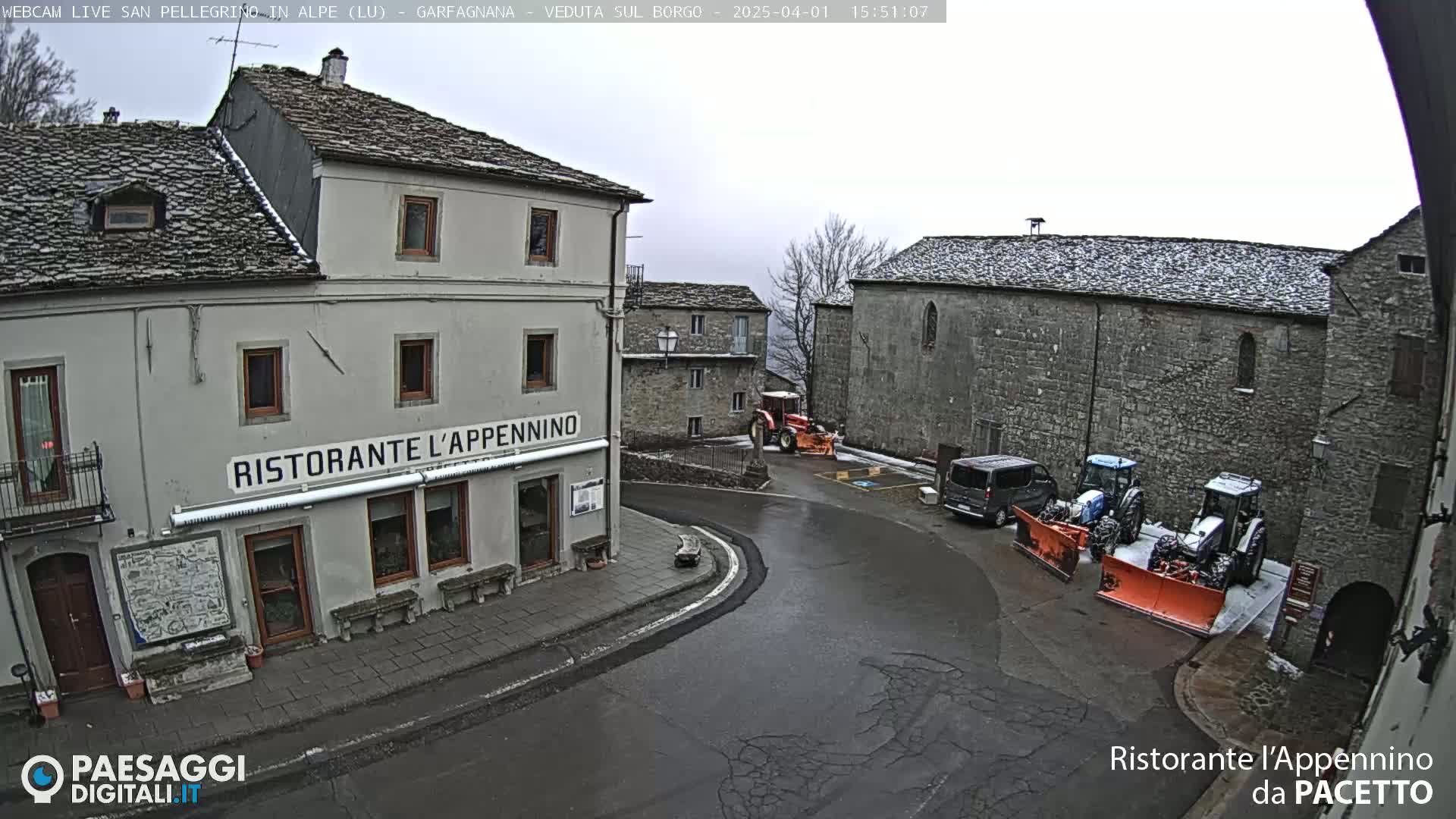 A snow-covered village square with several buildings, including a restaurant, and two tractors with snow plows parked nearby.