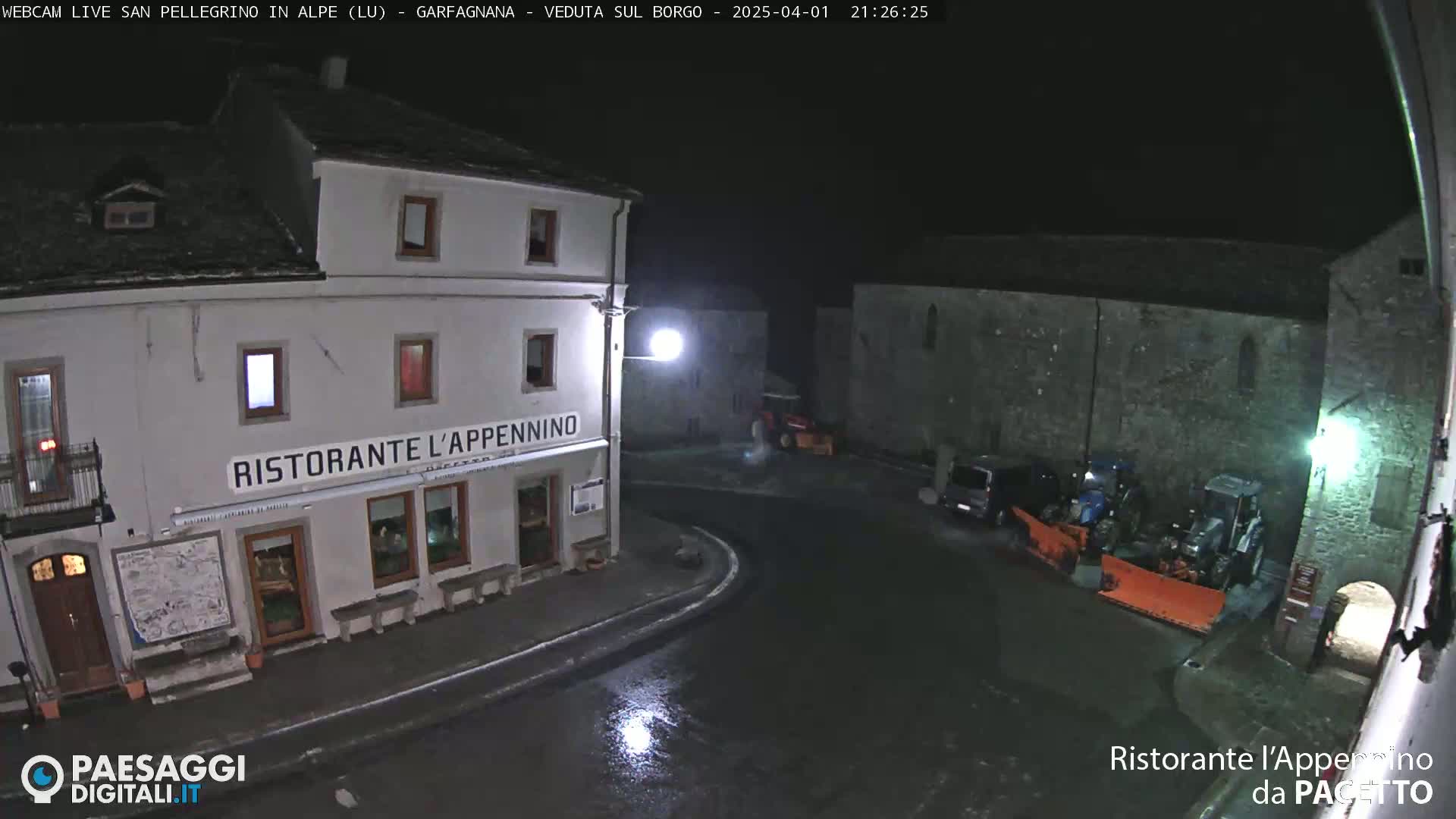 A nighttime view of a town square with a restaurant, some parked vehicles, and snow-removal equipment, under wet conditions.