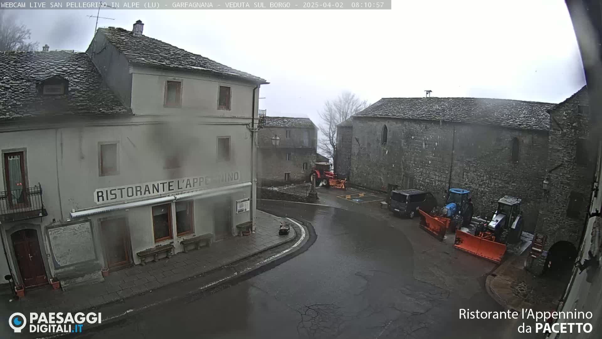 A rain-swept village square, featuring stone buildings, a restaurant, and several pieces of snow-clearing equipment.