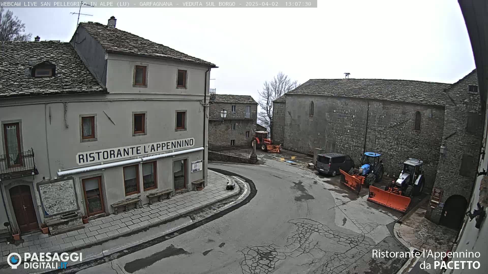 A gray stone village square with several buildings, two snow plows, and a van, under an overcast sky.