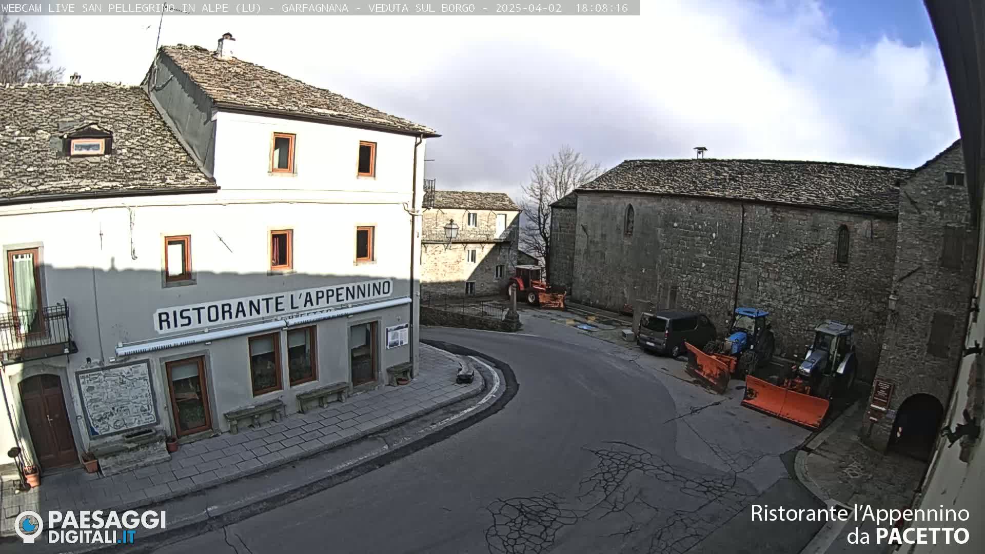 A village square with stone buildings, a restaurant, and several tractors with snowplows under a partly cloudy sky.