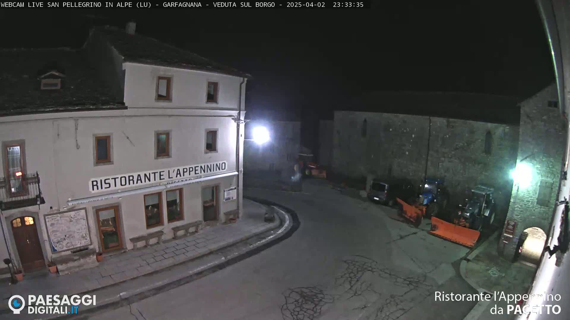 A nighttime, snow-free view of a town square with a restaurant, a few parked vehicles, and some snow removal equipment.