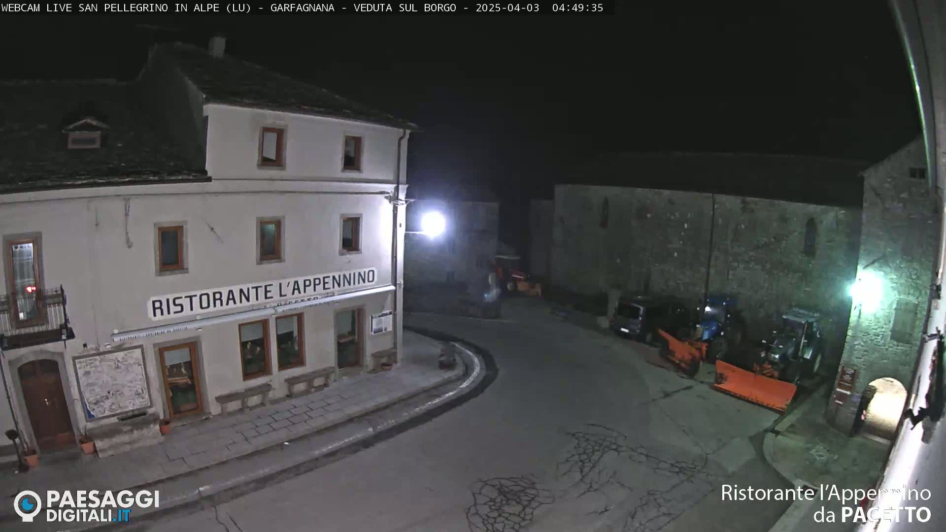 A nighttime, high-angle view of a town square with a restaurant, some parked vehicles, and snow removal equipment.