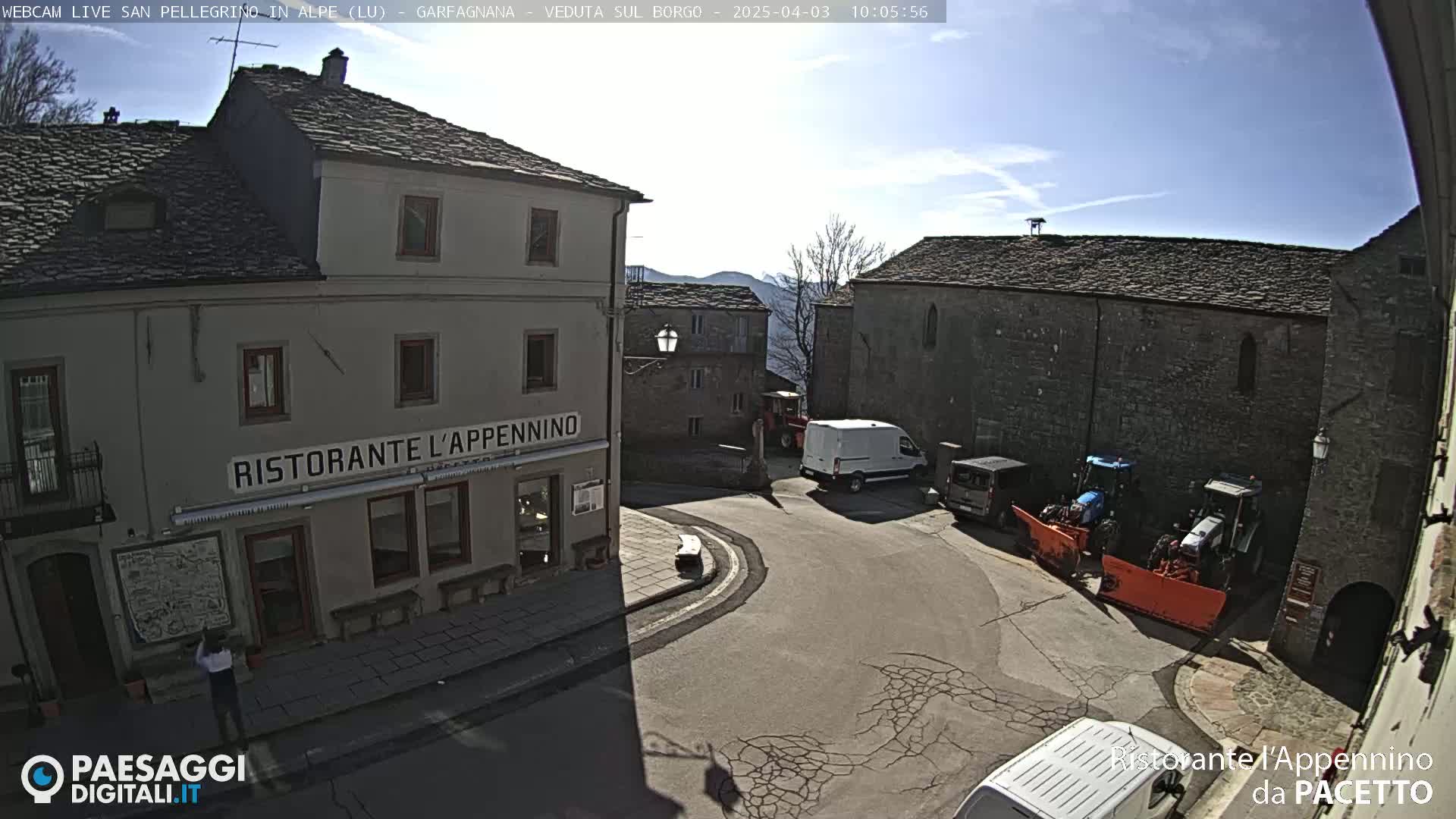 A sunny village square with stone buildings, a restaurant, a few vehicles, and tractors with snowplows.