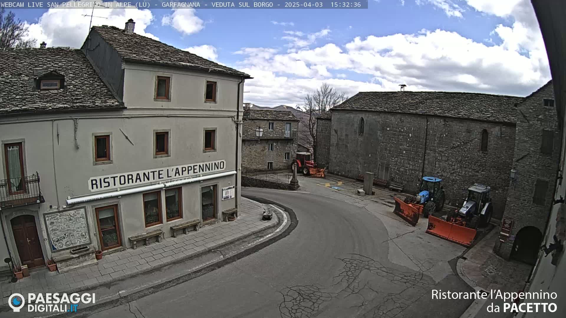 A partly cloudy day reveals a village square with stone buildings, including a restaurant, and two tractors with snowplows parked nearby.