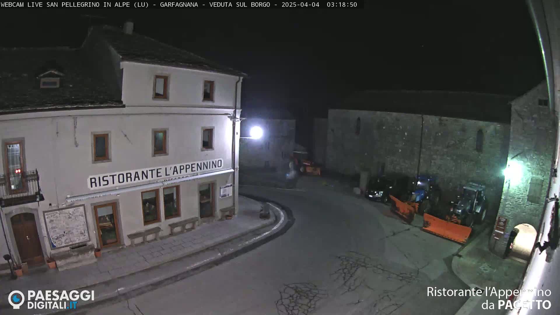 At night, a snowplow sits in a small town square next to a restaurant, with other vehicles parked nearby.