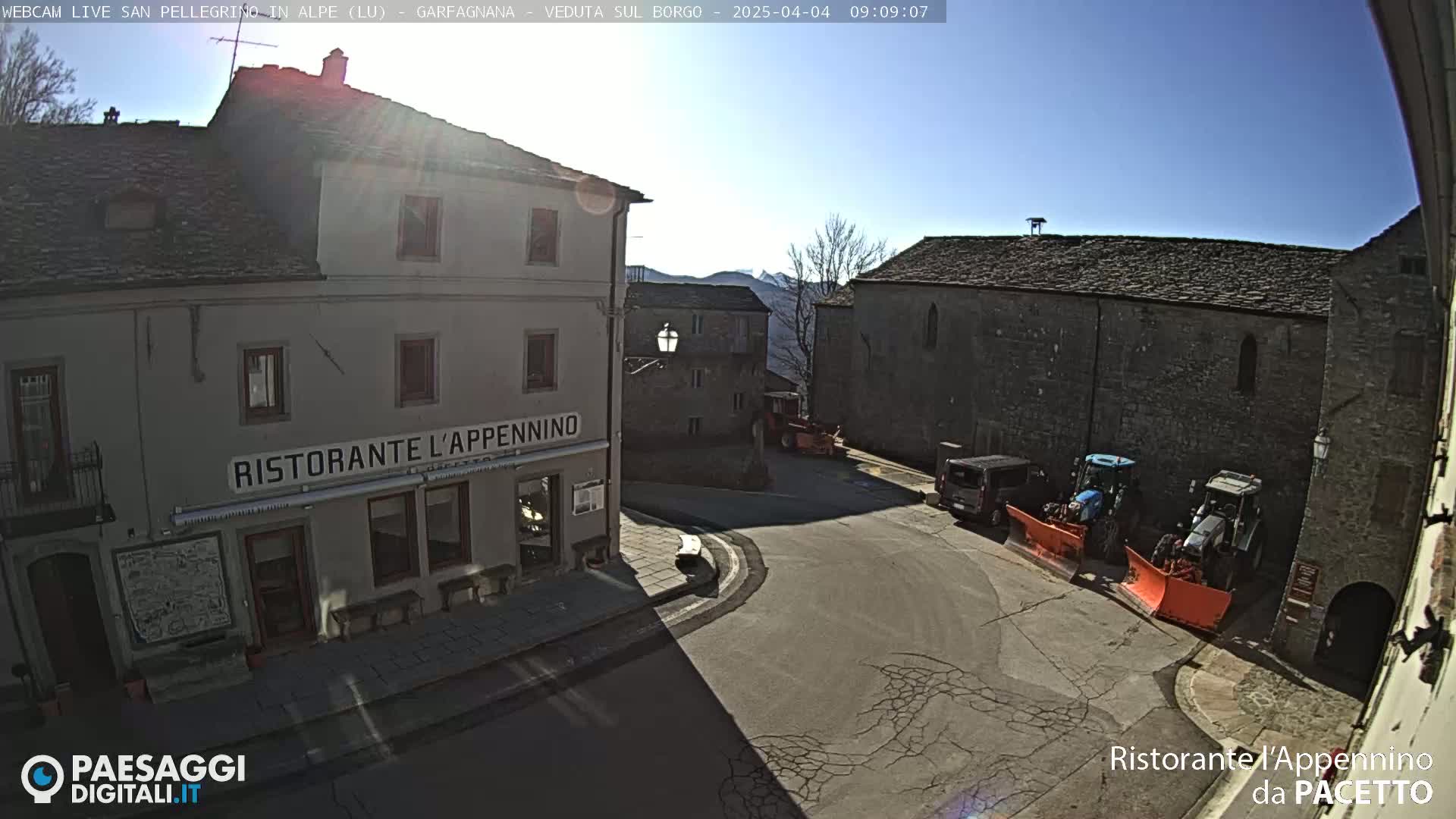 A sunny, clear day reveals a town square with stone buildings, a restaurant, and snow plows parked near a large stone structure.