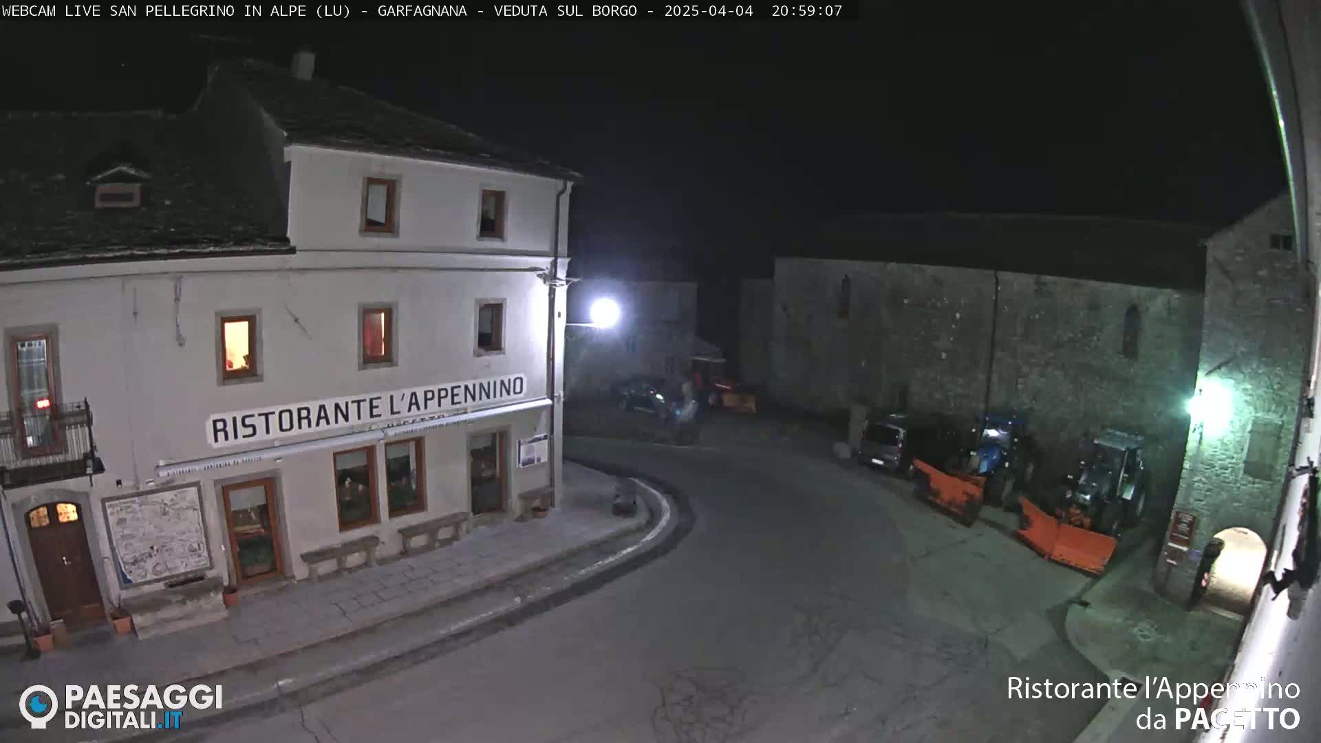 A nighttime, snow-covered village square scene features a restaurant called Ristorante L'Appennino and several parked vehicles, including snowplows.