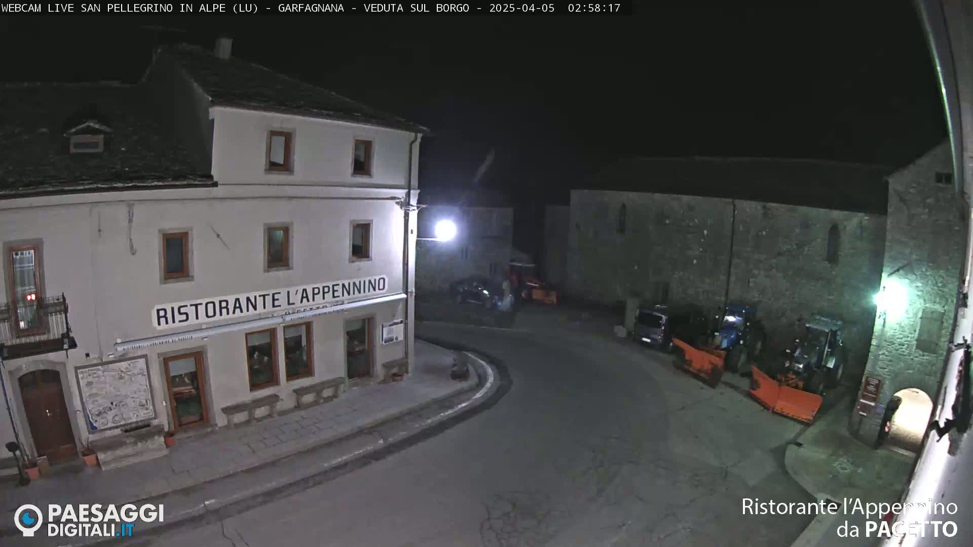 A nighttime view of a town square with a restaurant, several parked vehicles, and snowplows.