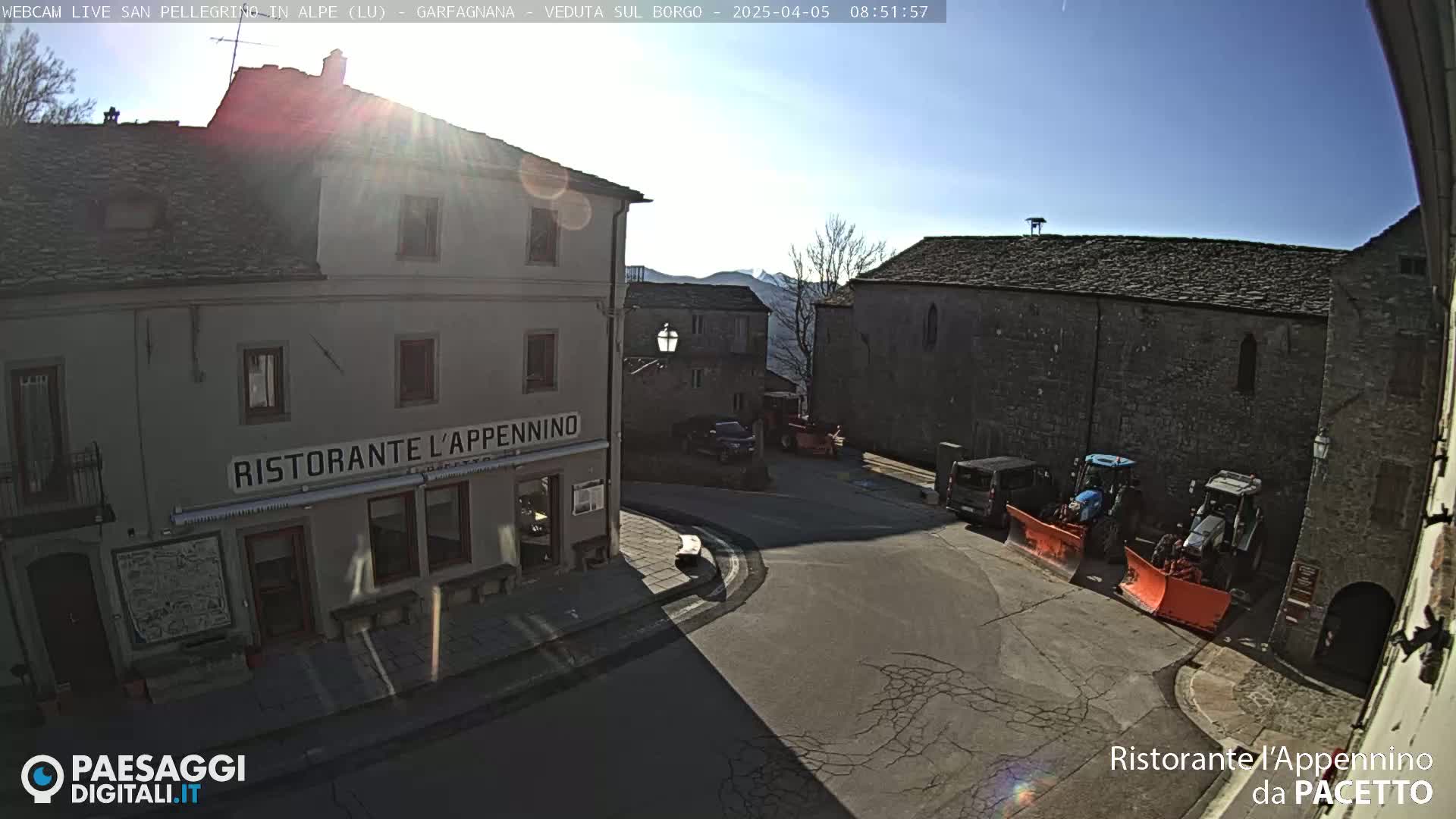 A sunny, snow-free village square with stone buildings, a restaurant, parked cars, and snowplows.