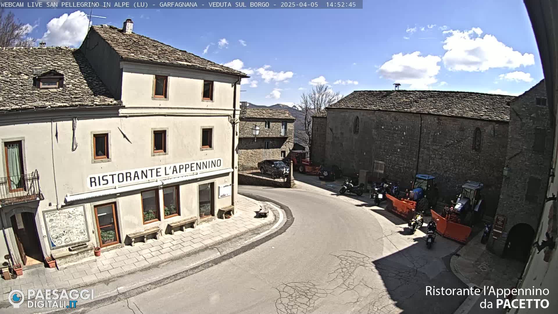 A sunny day shows a quaint village square with stone buildings, parked vehicles, and motorcycles, one building displaying the name of a restaurant.