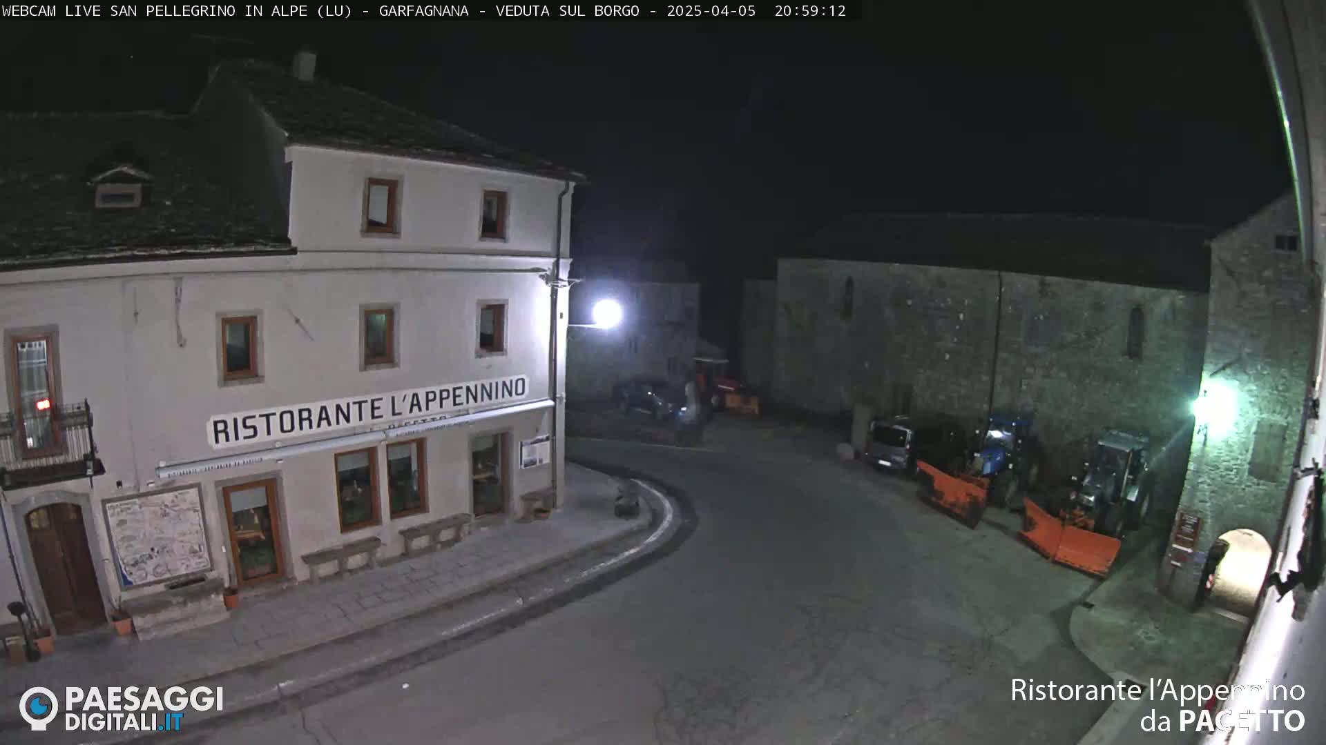 A nighttime view of a town square with a restaurant, stone buildings, and snow-clearing vehicles parked nearby under a clear, dark sky.