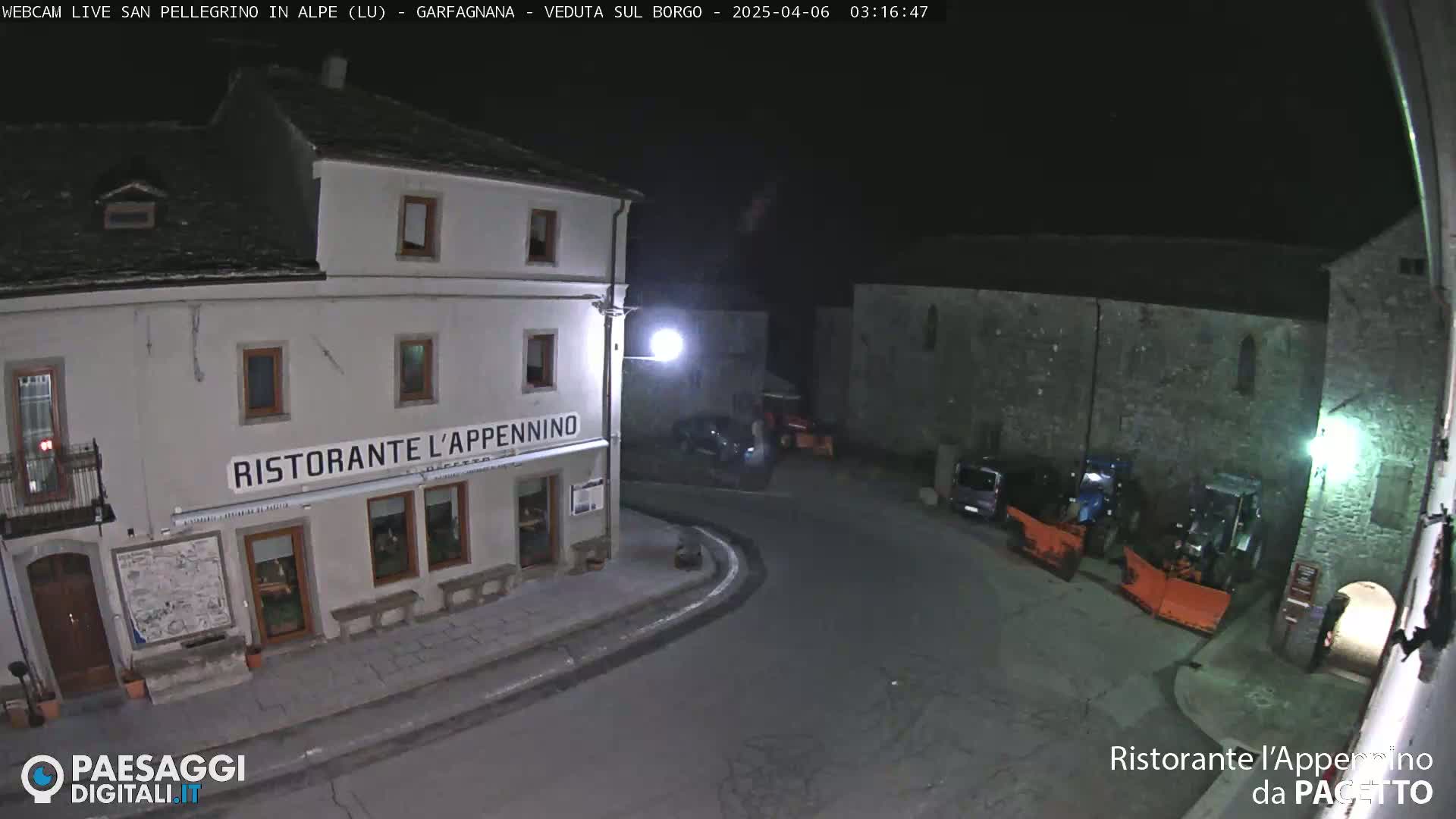 A nighttime view of a town square with a restaurant, several parked vehicles, and snow-removal equipment.