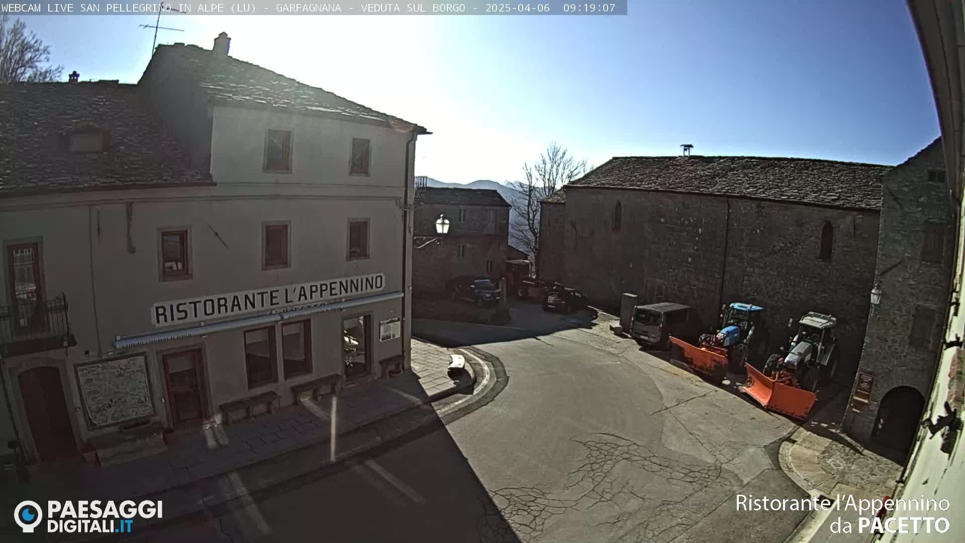 A sunny view of a town square with stone buildings, parked vehicles, and snowplows.