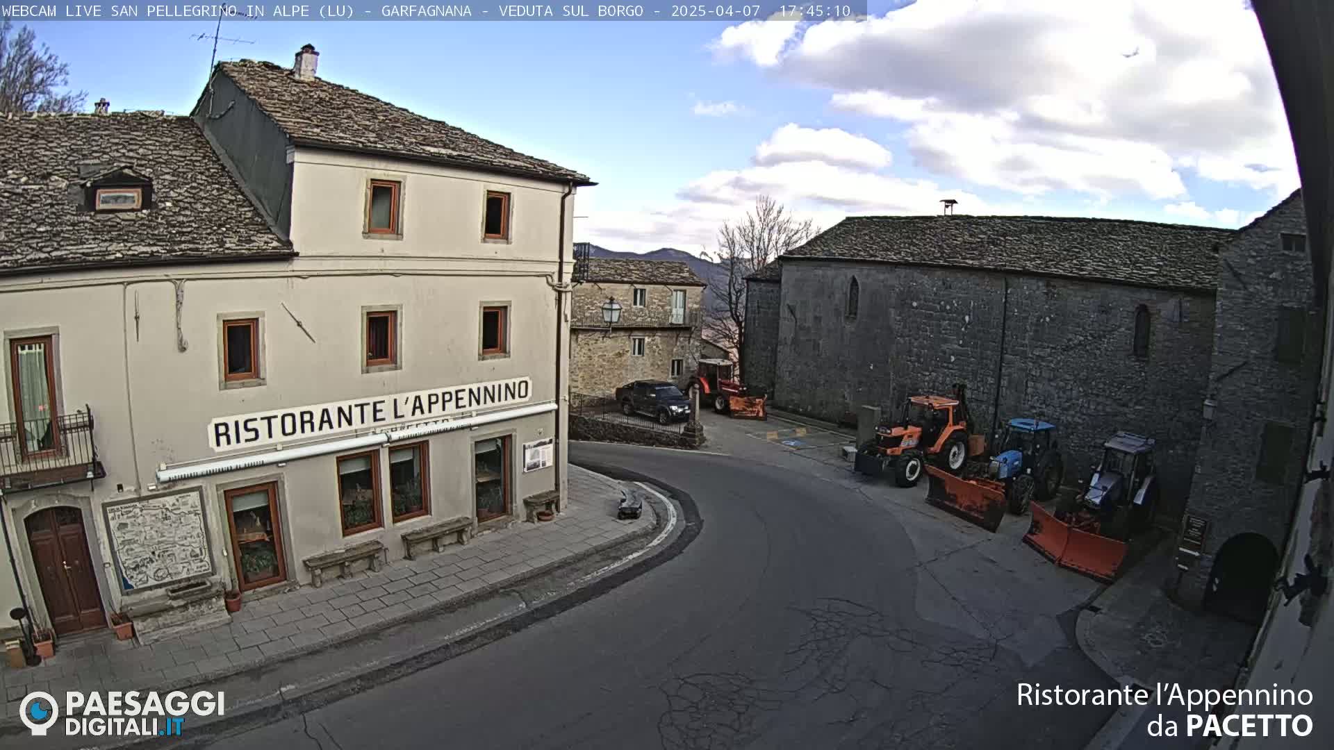 A partly cloudy day shows a street corner in a town with stone buildings, one of which is a restaurant, and several tractors with snow plows parked nearby.