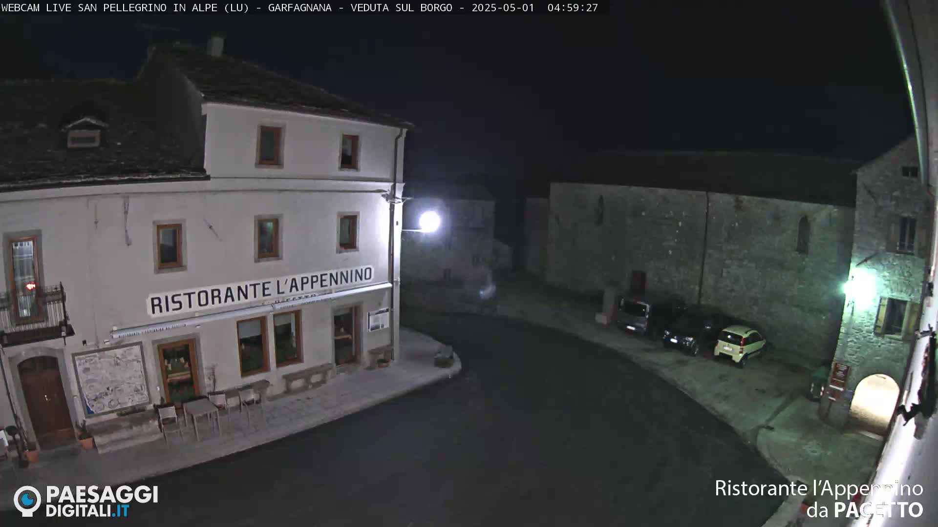 At night, a village square with several parked cars is illuminated by streetlights, featuring a restaurant called "Ristorante l'Appennino."