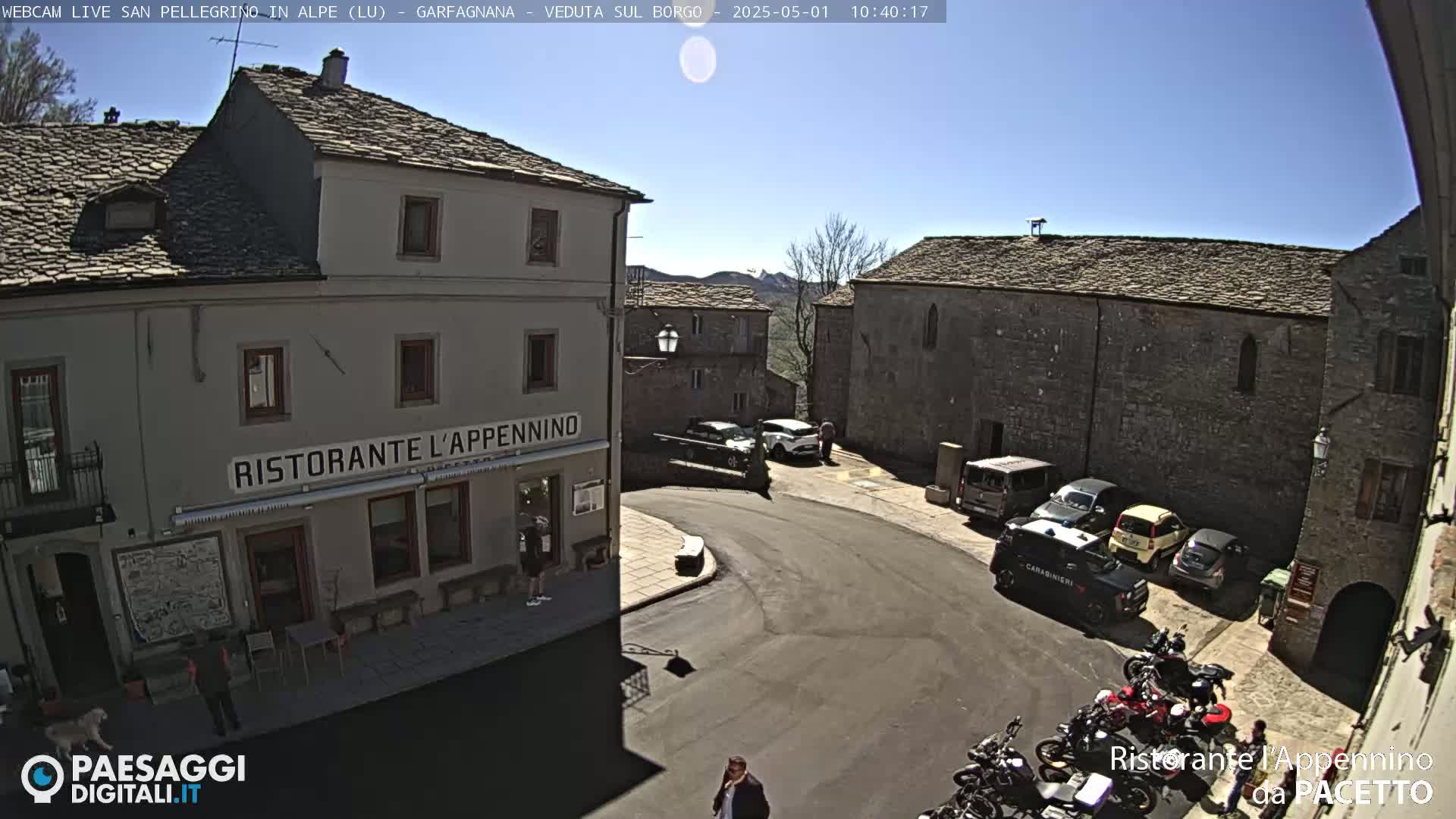 A sunny view of a town square with several parked cars and motorcycles, stone buildings, and a restaurant.