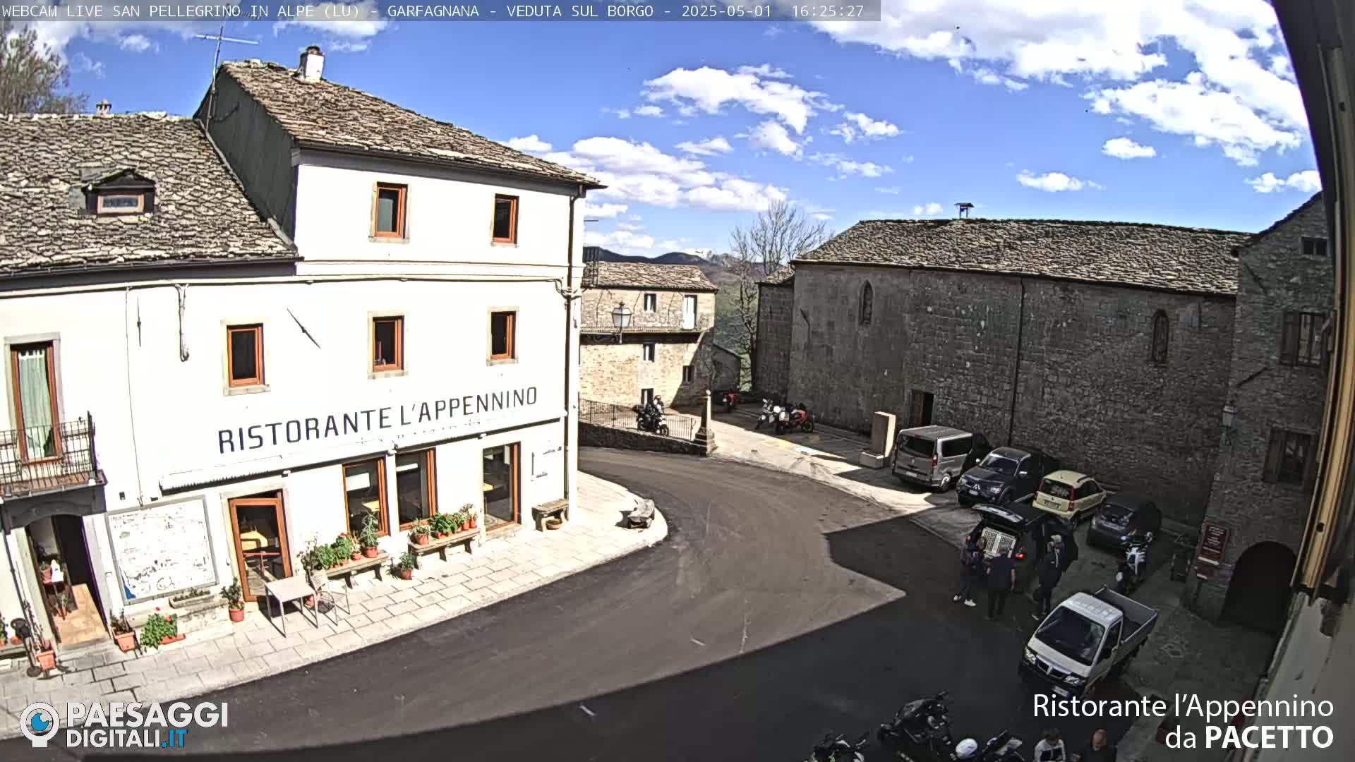 A sunny day reveals a quaint village square with stone buildings, a restaurant, parked cars, and several motorcycles, under a partly cloudy sky.