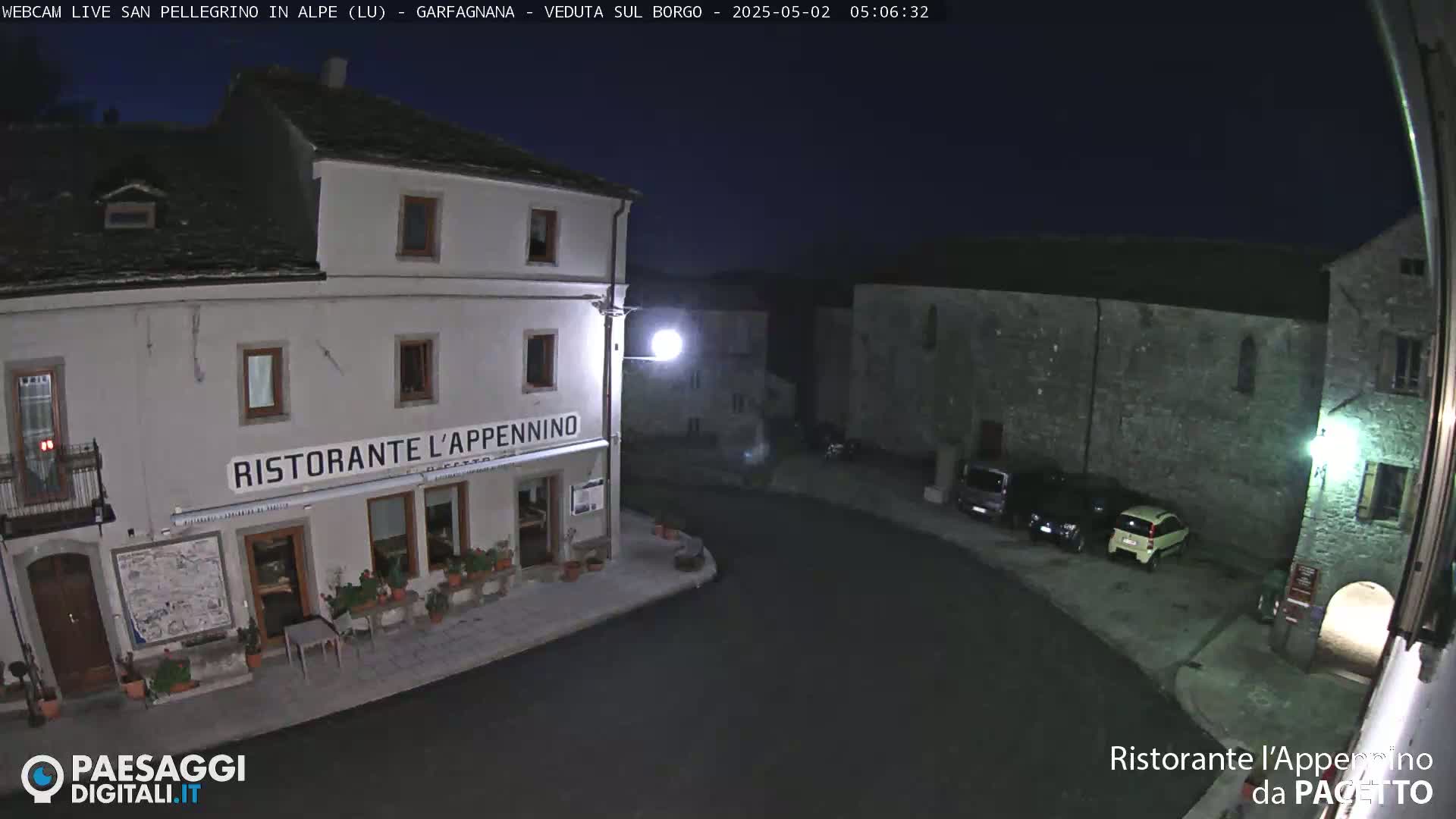 A nighttime view of a village square with a restaurant, several parked cars, and stone buildings under a dark sky.