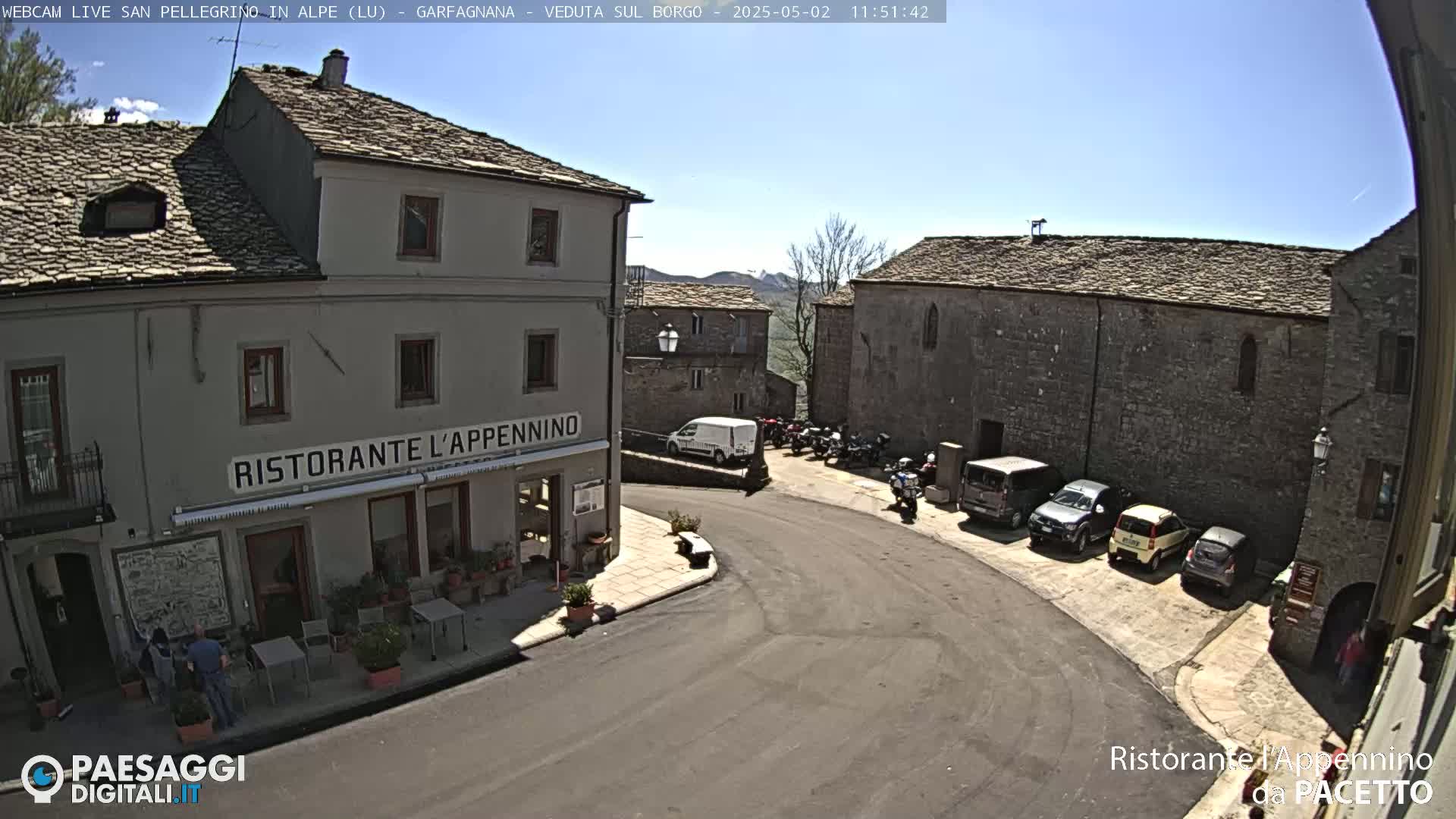 A sunny day reveals a village square with several parked cars and motorcycles surrounding a restaurant.