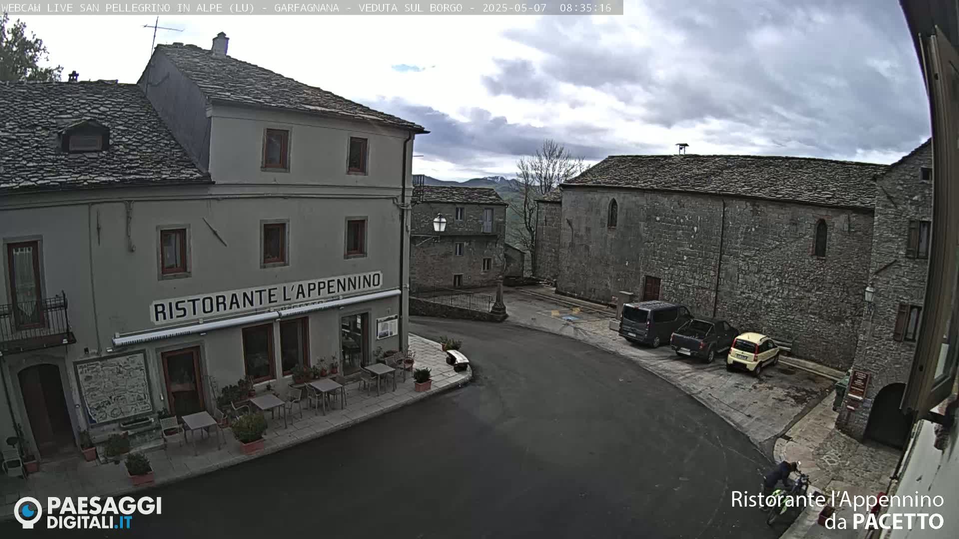 A stone village square under a cloudy sky features a restaurant with outdoor seating, parked vehicles, and a cyclist.