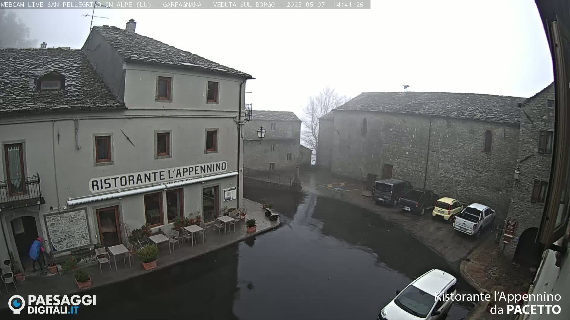 A foggy village square with stone buildings, a restaurant with outdoor seating, and several parked cars is partially flooded.