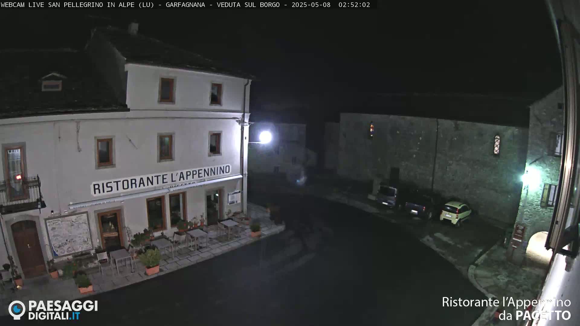 A nighttime view of a town square with a restaurant, several parked cars, and stone buildings under a dark sky.
