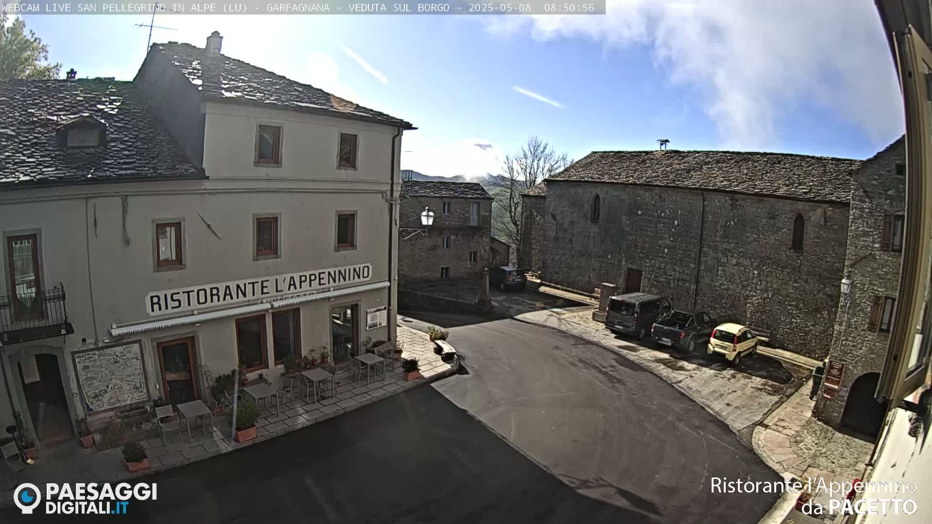 A partly cloudy day reveals a village square with several stone buildings, a restaurant with outdoor seating, and a few parked cars.