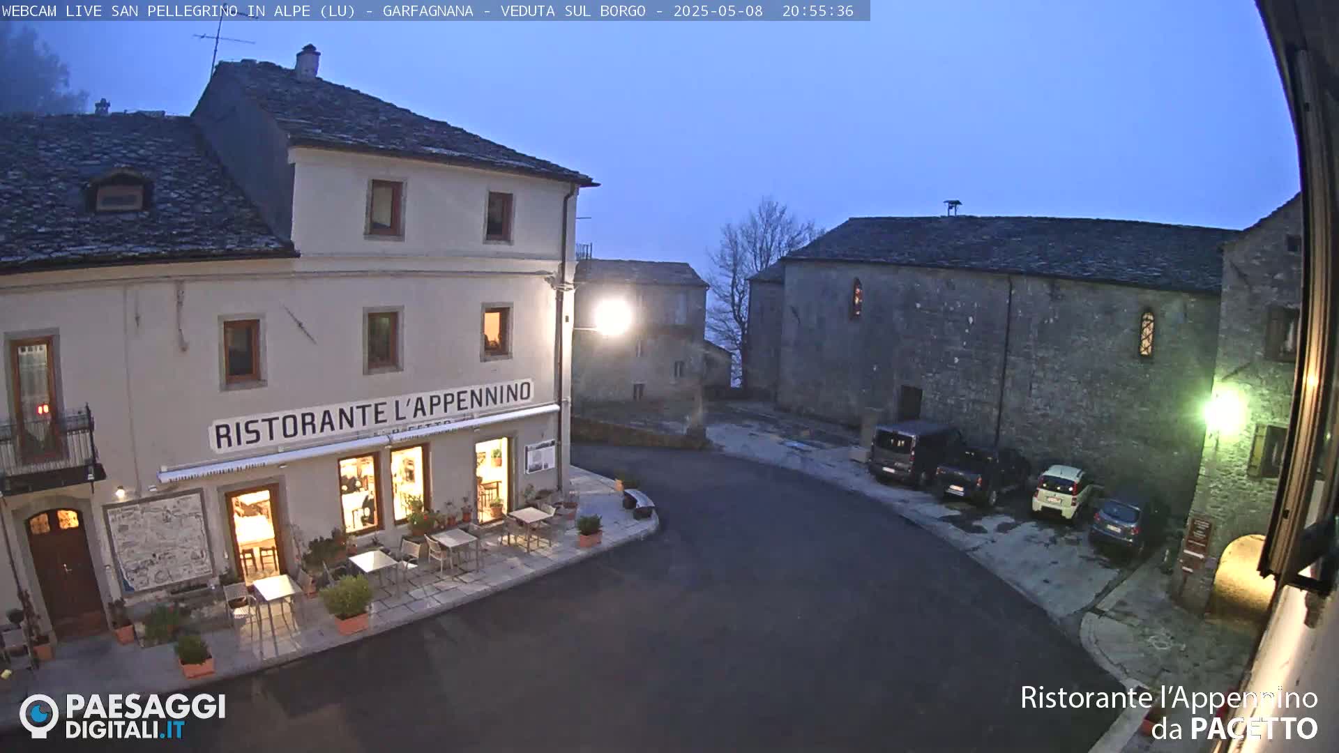 A dimly lit village square at twilight, featuring a restaurant with outdoor seating and several parked cars, under a clear, dark sky.