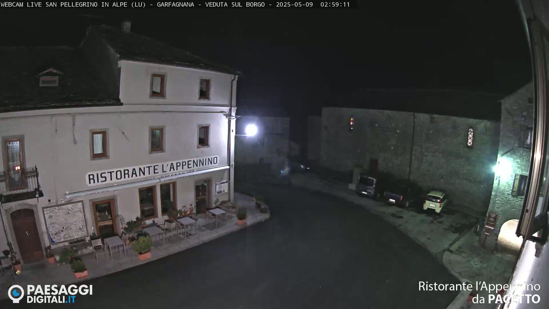 A nighttime view of a town square with a restaurant and several parked cars under a clear, dark sky.