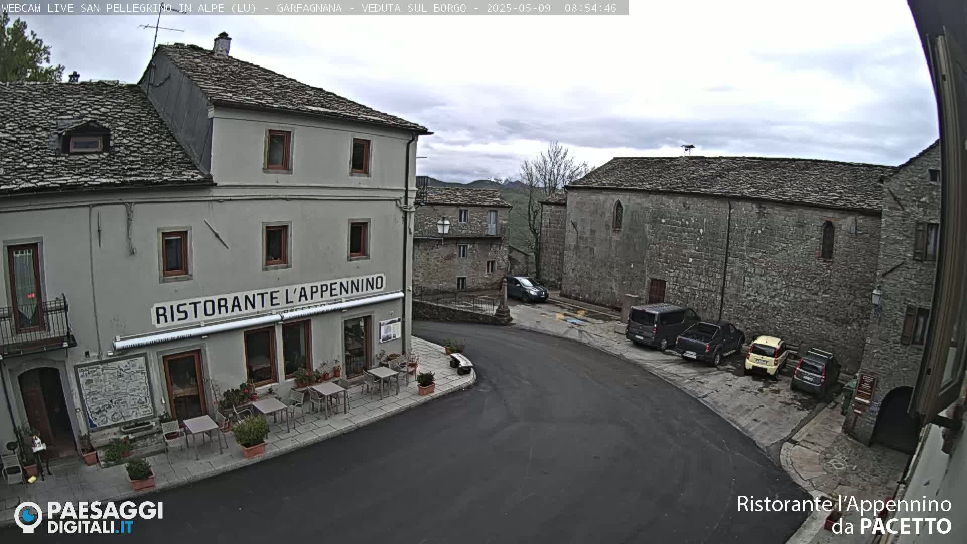 A street-level view of a village square on an overcast day, showing several stone buildings, parked cars, and outdoor seating at a restaurant.