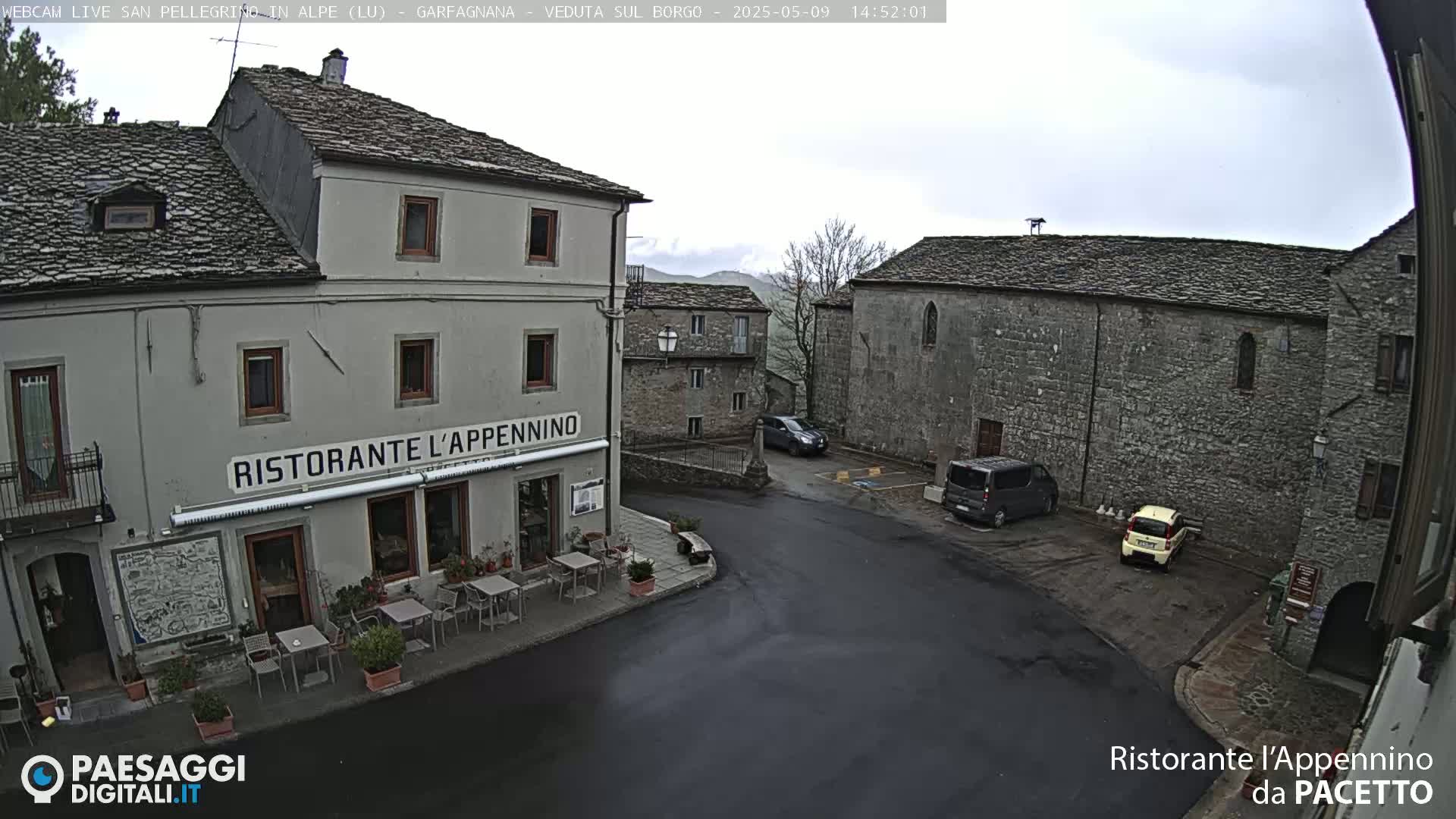 A town square with stone buildings, a restaurant with outdoor seating, and a few parked cars under an overcast sky.