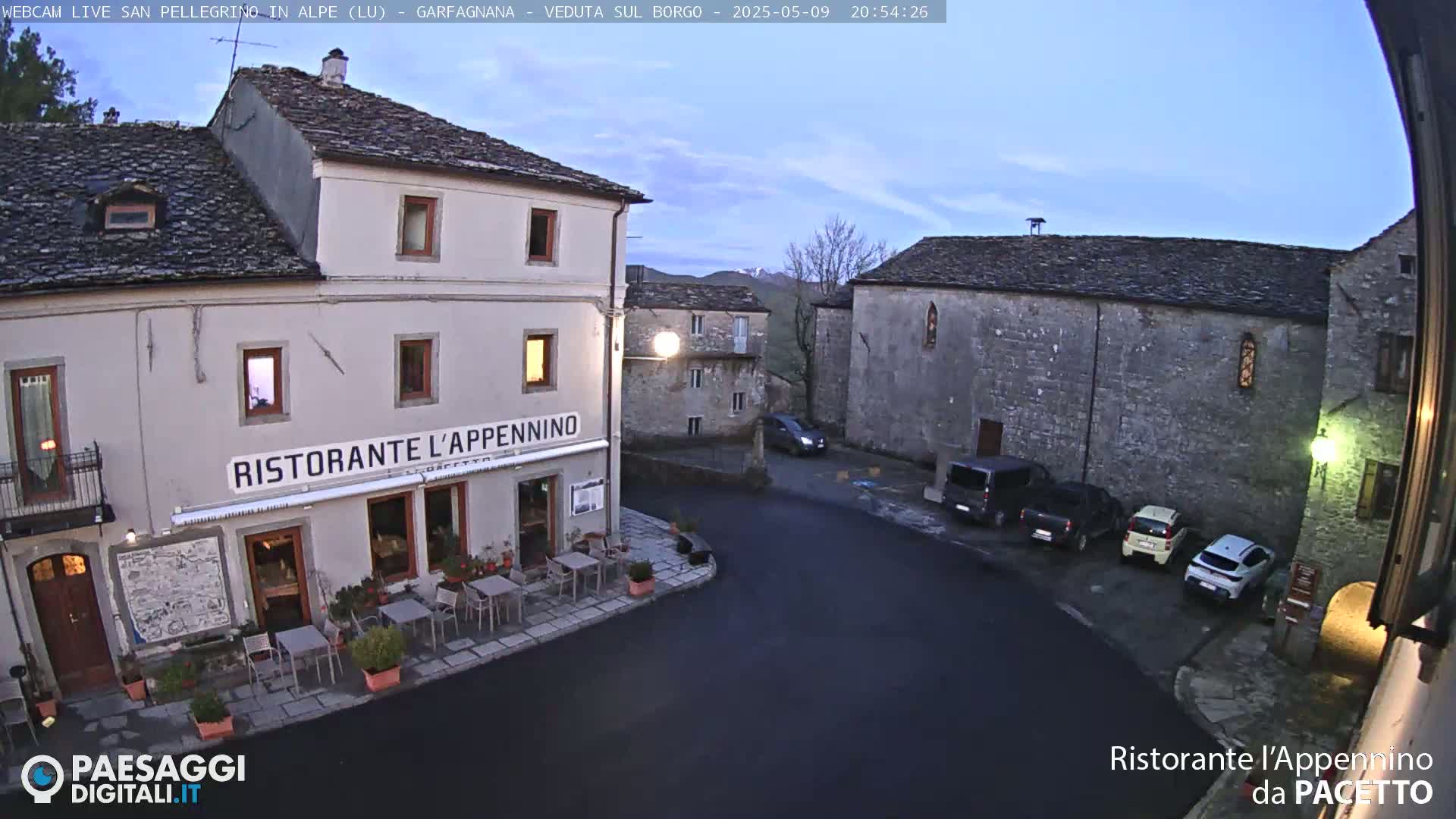 A village square at dusk, under a clear sky, shows several parked cars near a restaurant with outdoor seating.