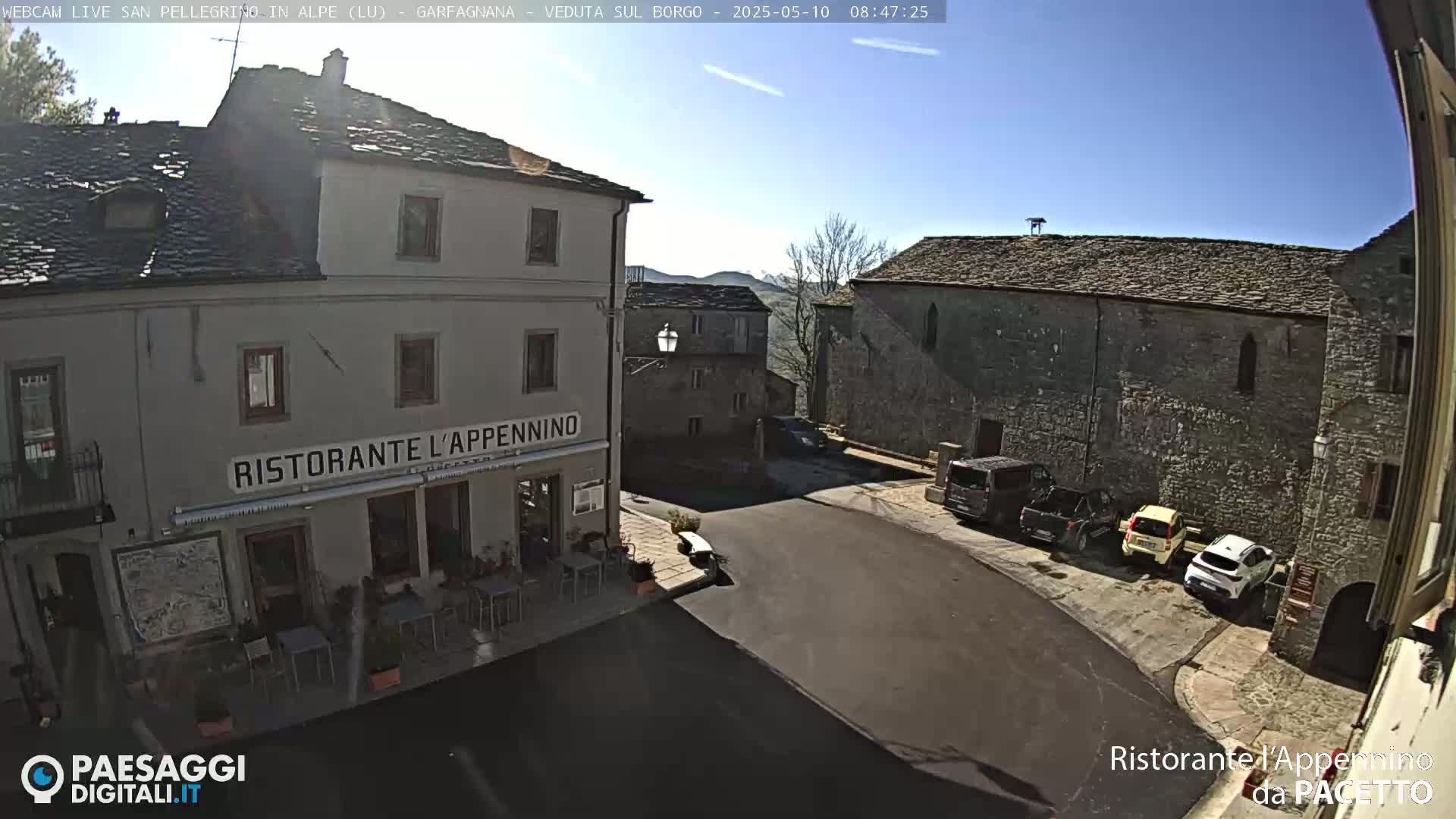 A sunny, outdoor view of a town square with several parked cars, a restaurant, and stone buildings.
