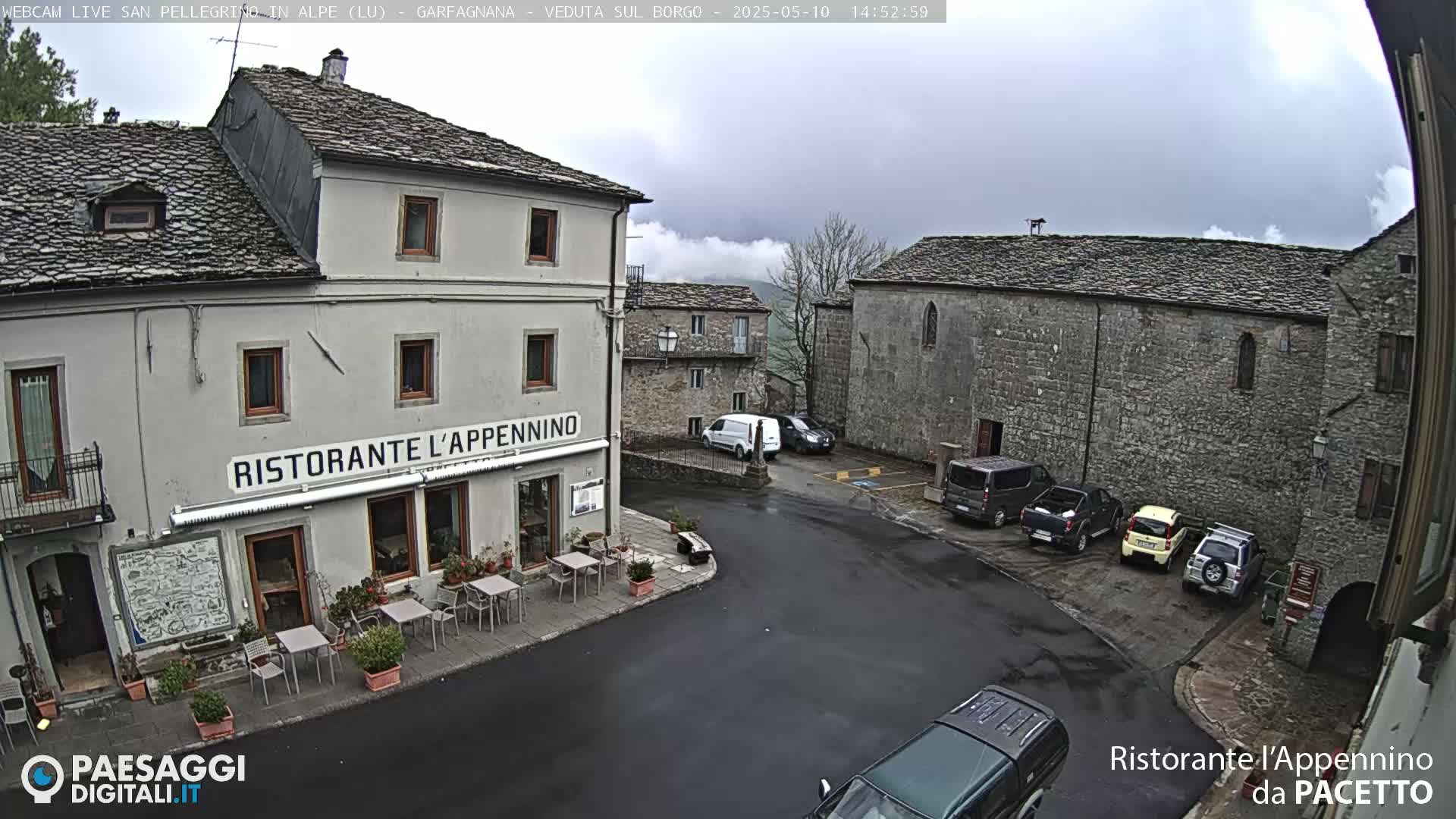 A restaurant sits in a town square under an overcast sky, with several cars parked nearby and stone buildings surrounding the square.